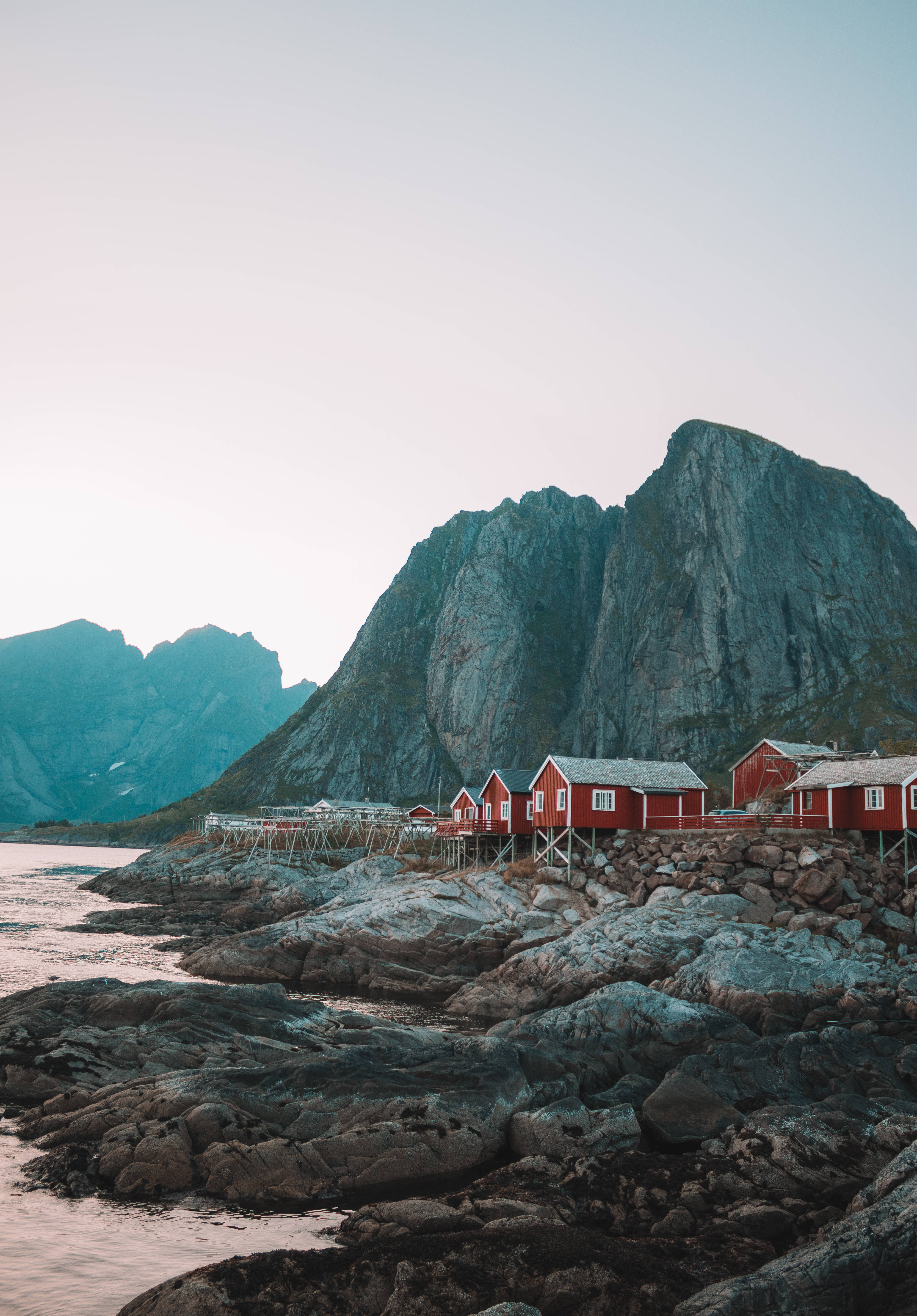 Norway 桌布 壁紙 — 紅色木屋坐落在岩石海岸上，旁邊是高聳的灰色山脈，天空呈現柔和的暮色。