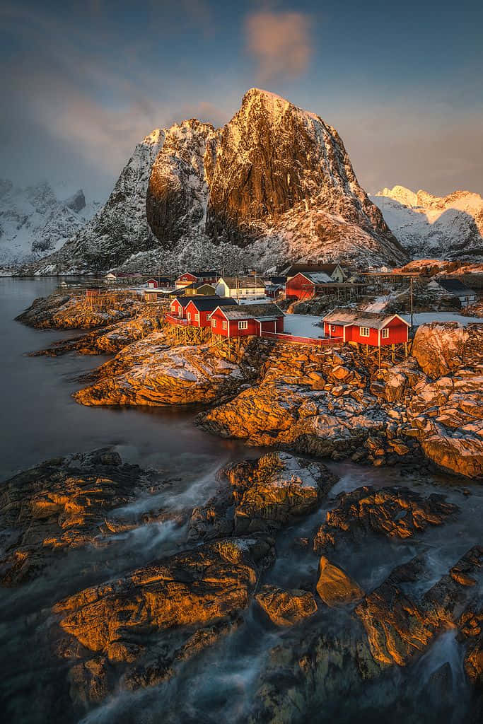 Winter Photography 桌布 壁紙 — 紅色木屋坐落在白雪覆蓋的岩石海岸上，下方是沐浴在金色日落光芒中的高聳山脈。