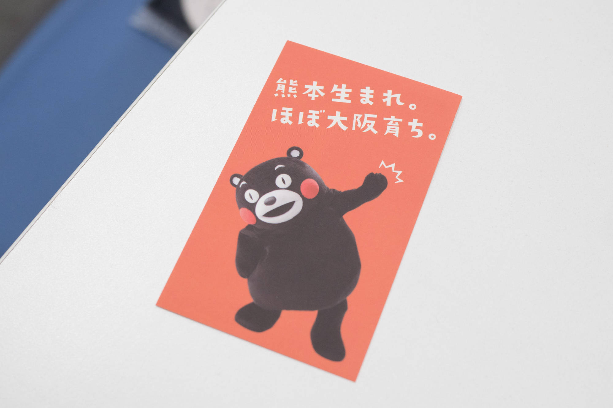 紅色熊本熊宣傳單 壁紙 — 一張帶有日文字樣的開心 Kumamon 貼紙，放在白色表面上，旁邊有一個藍色物件。