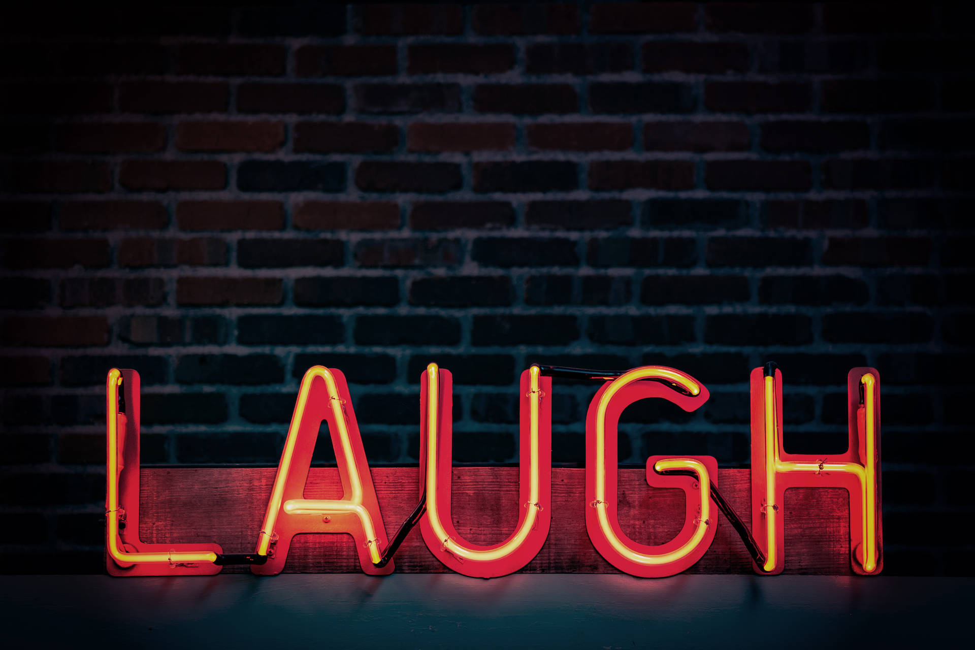 一個紅色霓虹燈牌，拼寫著 LAUGH，放在一塊木板上，靠著磚牆。