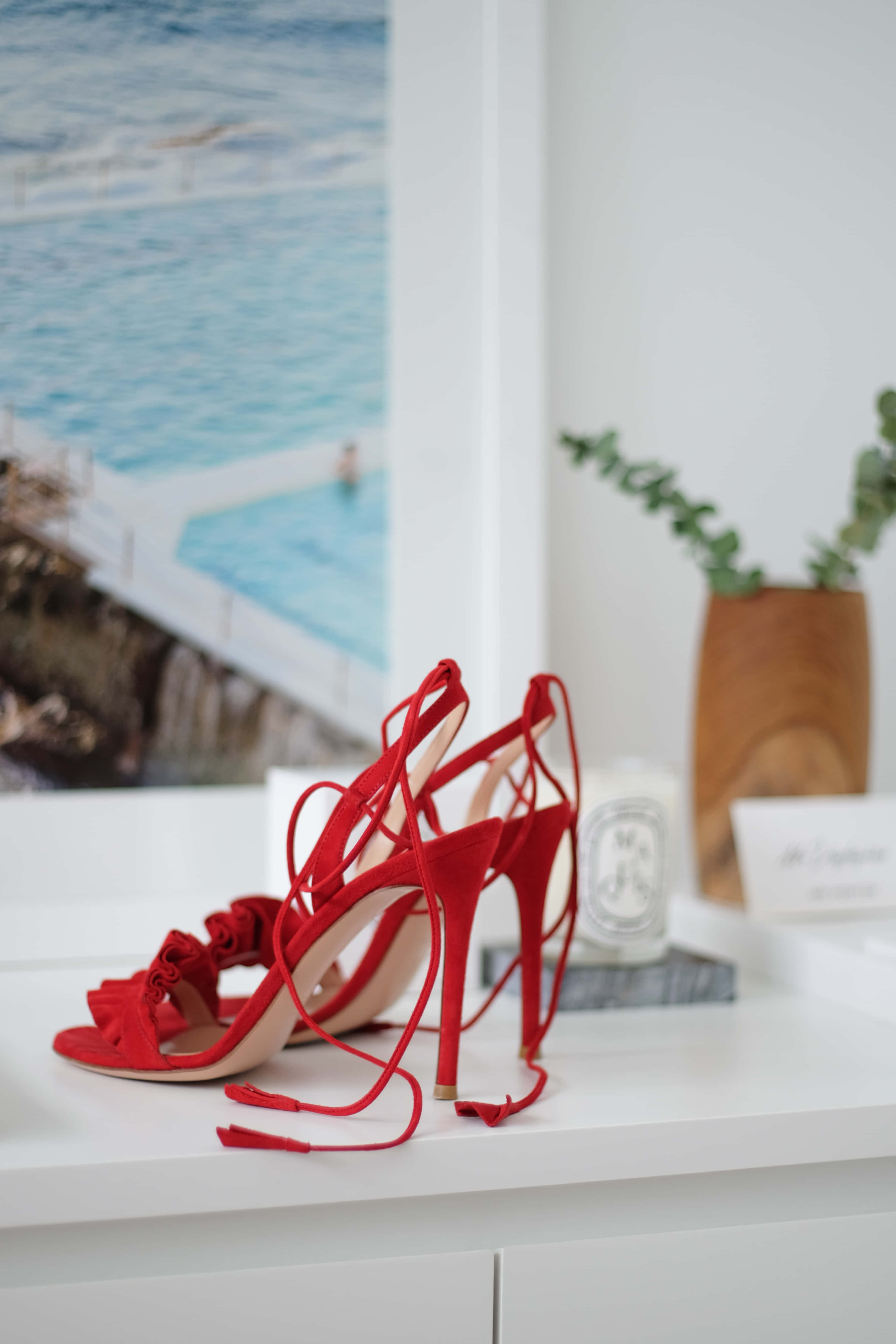 Red High Heels 桌布 壁紙 — 紅色綁帶高跟鞋放在白色架子上，旁邊有蠟燭和尤加利葉，背後是模糊的海景。