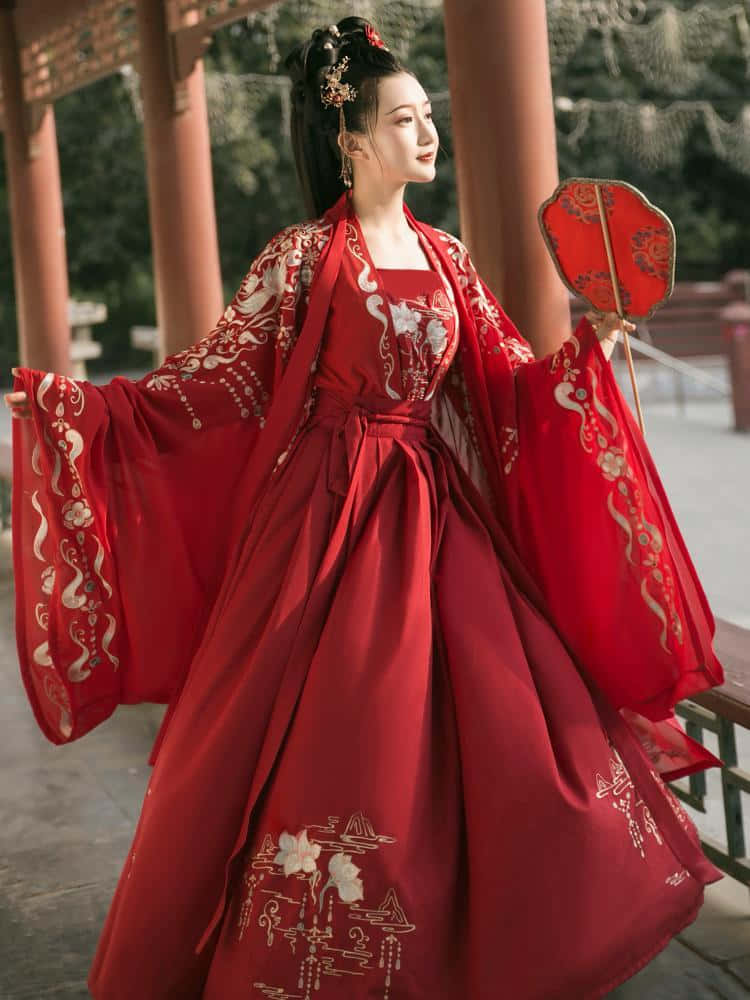 「紅色美學之美，在於其大膽而熱情的氣息。」 壁紙 — 一位身穿飄逸紅色傳統長袍、飾有金色刺繡的女子，優雅地站在木亭下，手持相配的扇子。