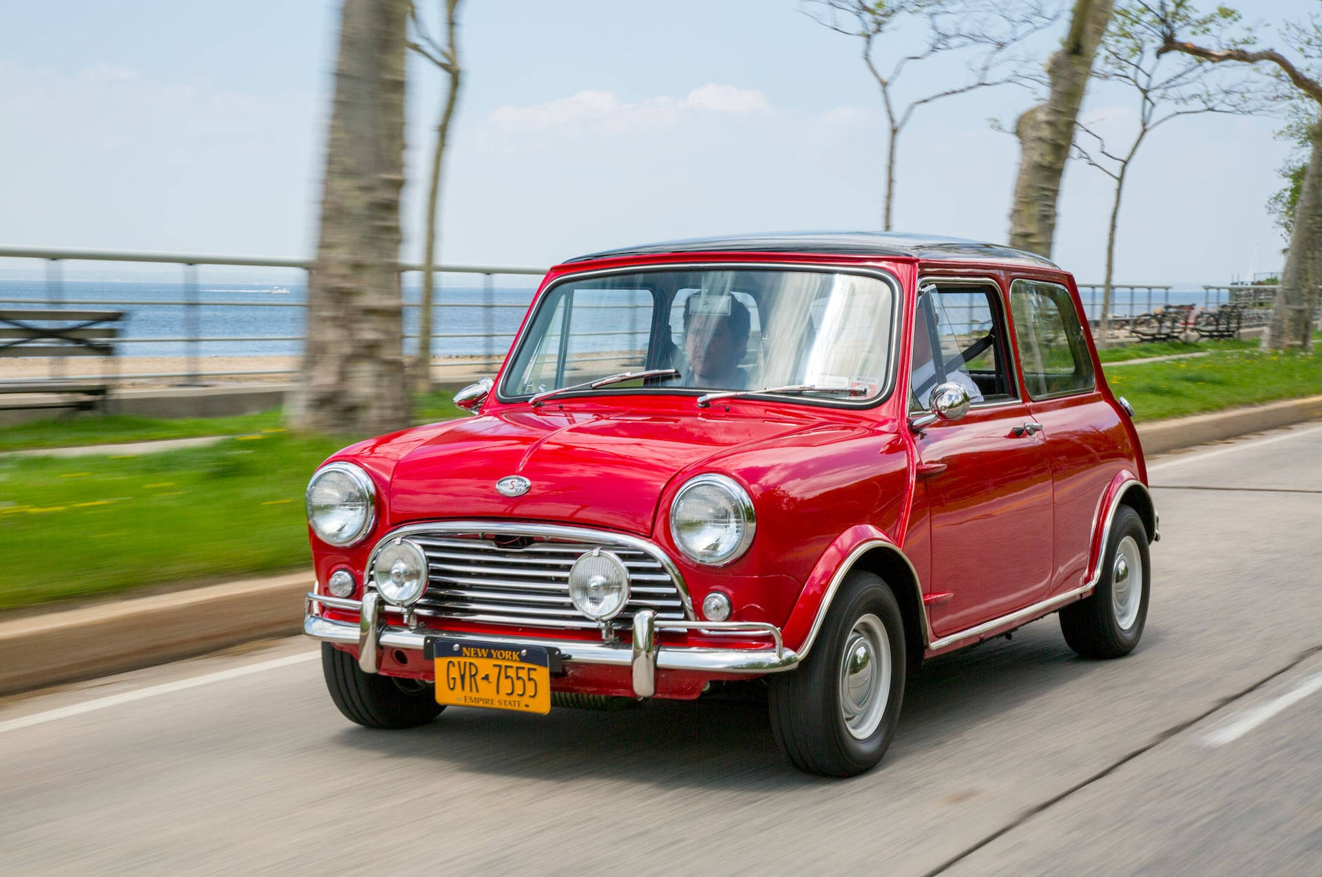 紅色美學迷你汽車行駛於道路 壁紙 — 一輛鮮紅色經典Mini Cooper沿海岸道路行駛，背景有模糊的樹木和海洋。