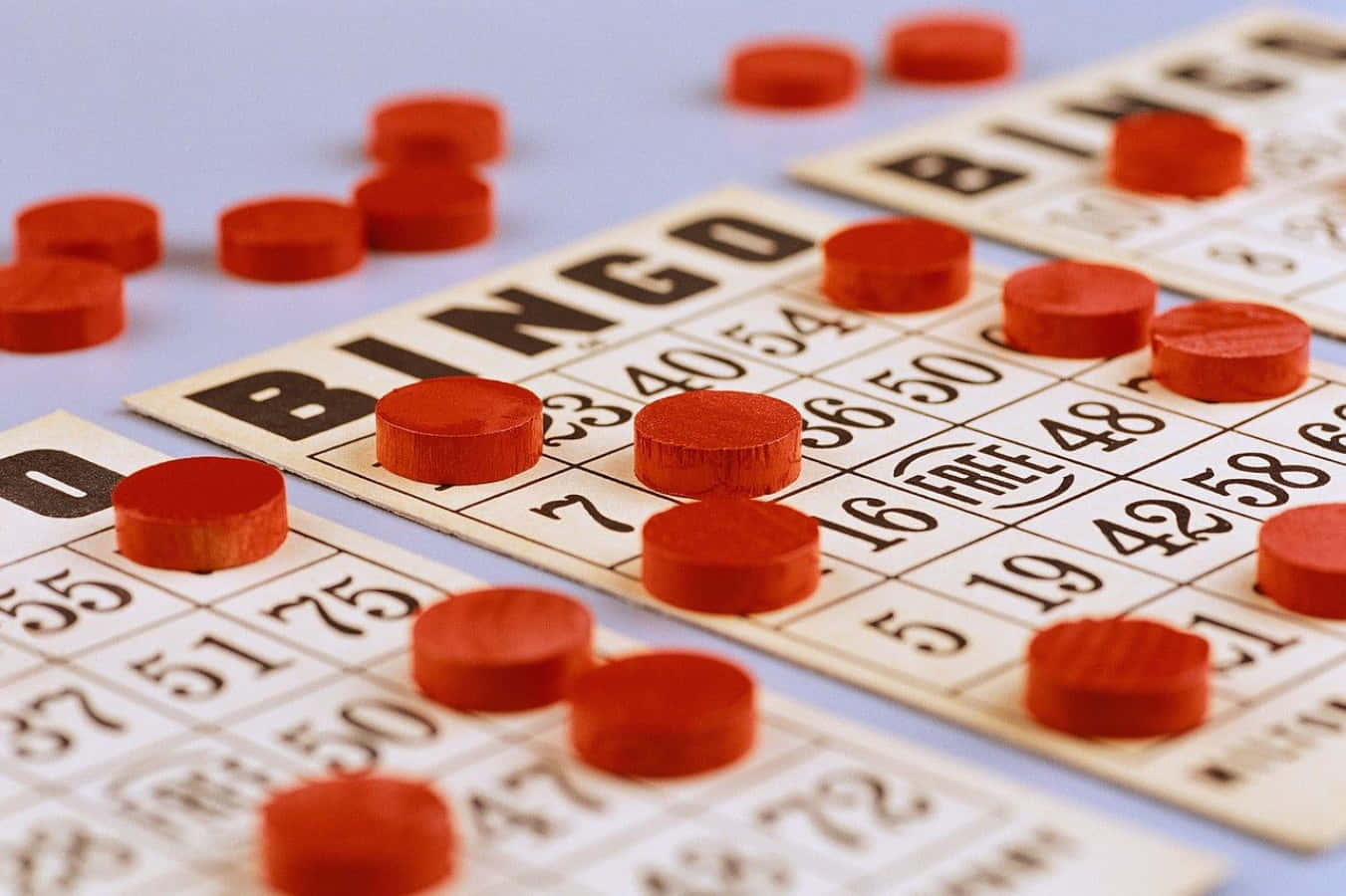 Bingo 桌布 壁紙 — 紅色賓果籌碼散落在多張印有粗體黑色數字的米色紙賓果卡上的特寫。