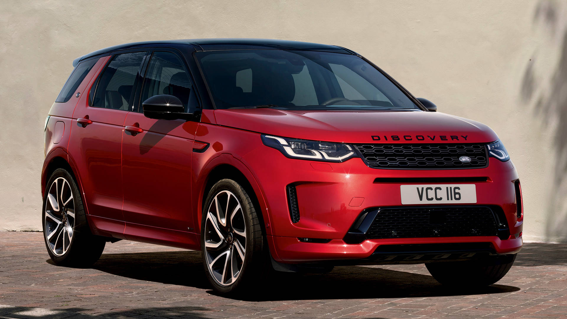 紅色路虎 iPhone 主題壁紙 壁紙 — 一輛亮紅色的 Land Rover Discovery SUV 停泊在磚砌路面上，背景是純色米色牆壁，在明亮日光下。