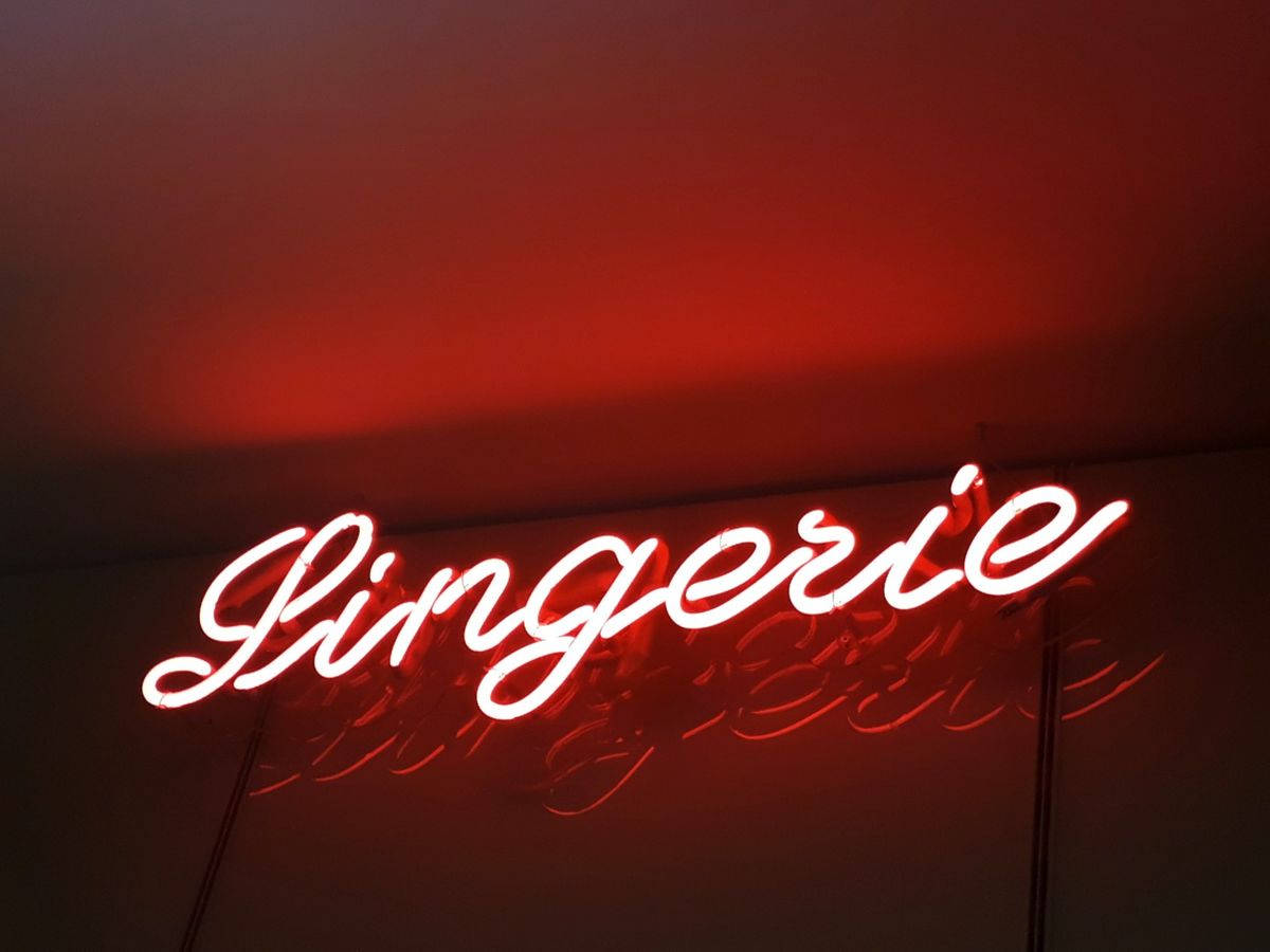 紅色霓虹風「Lingerie」招牌 壁紙 — 一個發光的紅色霓虹燈牌，以草書拼寫「Lingerie」，在黑暗的天花板上投射出溫暖的環境光。