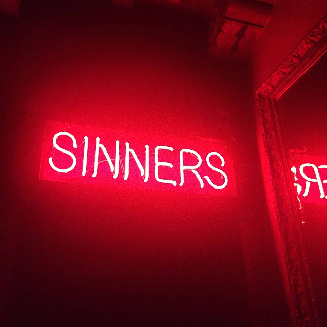 紅色霓虹風「罪人」 壁紙 — 一個發光的紅色霓虹燈牌，拼寫著 SINNERS，在昏暗的室內環境中，將強烈的光芒投射在帶有華麗線條的深色牆壁上。