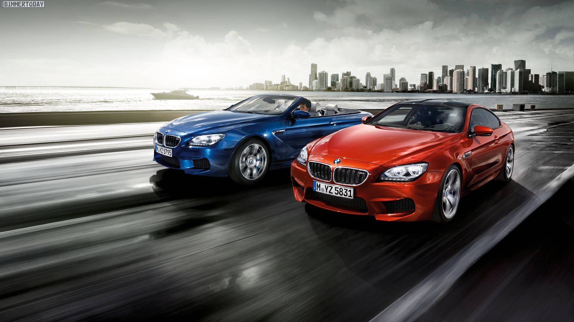紅藍BMW競速賽 壁紙 — 兩輛時尚的BMW跑車沿著濕漉漉的沿海高速公路奔馳，背景是現代化的城市天際線。