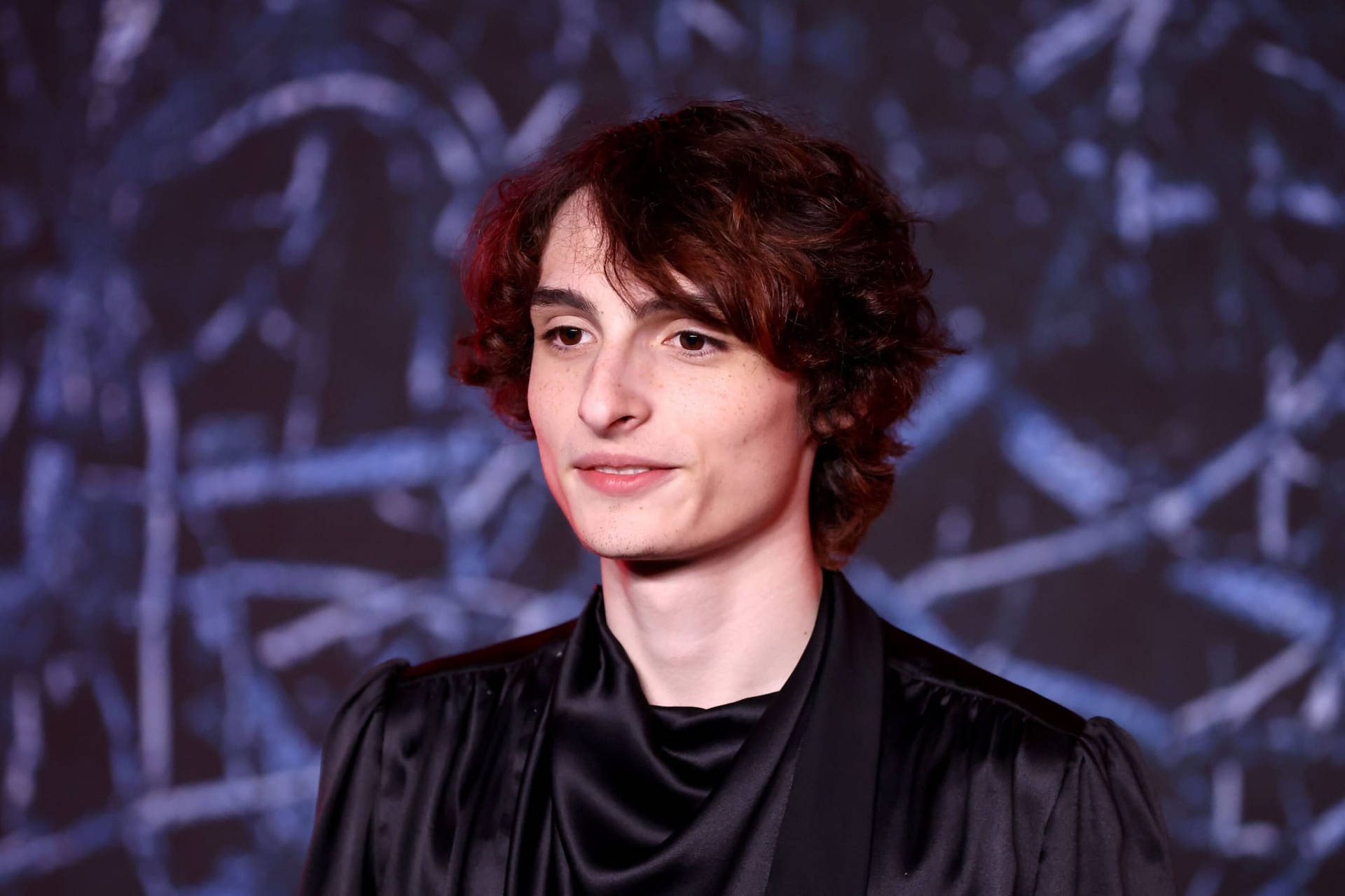 身穿黑色緞面長袍的年輕男子在抽象藍色背景前對著鏡頭微笑。