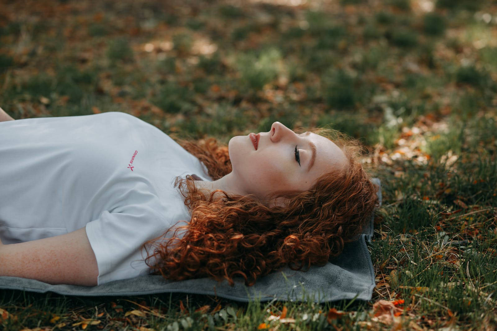 紅髮女子悠閒放鬆 壁紙 — 一位擁有紅色捲髮的年輕女性平靜地躺在散落著秋葉的草地上，閉著眼睛，享受寧靜的放鬆。