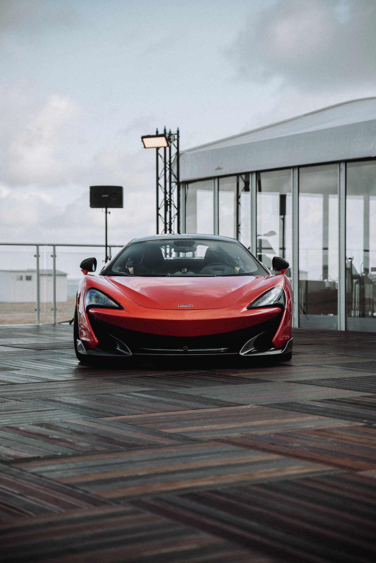 多雲天空下，現代玻璃建築附近木質甲板上的紅色McLaren跑車