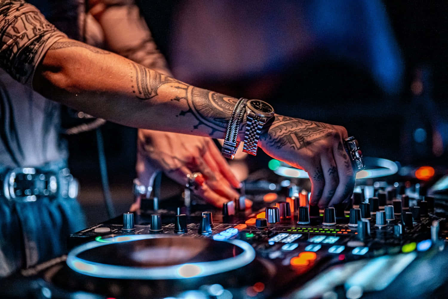 DJ 桌布 壁紙 — 紋身DJ雙手熟練地操控發光的旋鈕和唱盤，在充滿活力的夜店燈光下。