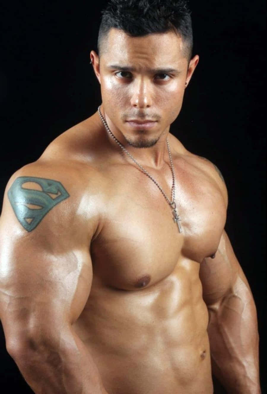 紋身肌肉男圖片 壁紙 — 一個肌肉發達嘅男人，肩膀上有一個超人紋身，喺黑色背景前強烈地凝視。