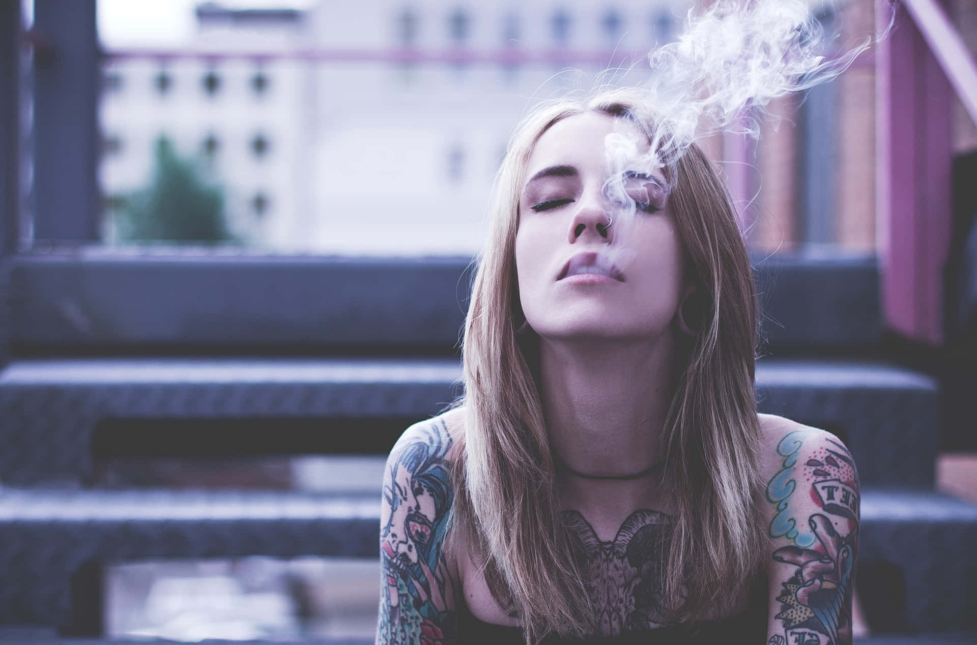 有紋身的女性坐在城市階梯上，閉著眼睛呼出煙霧。