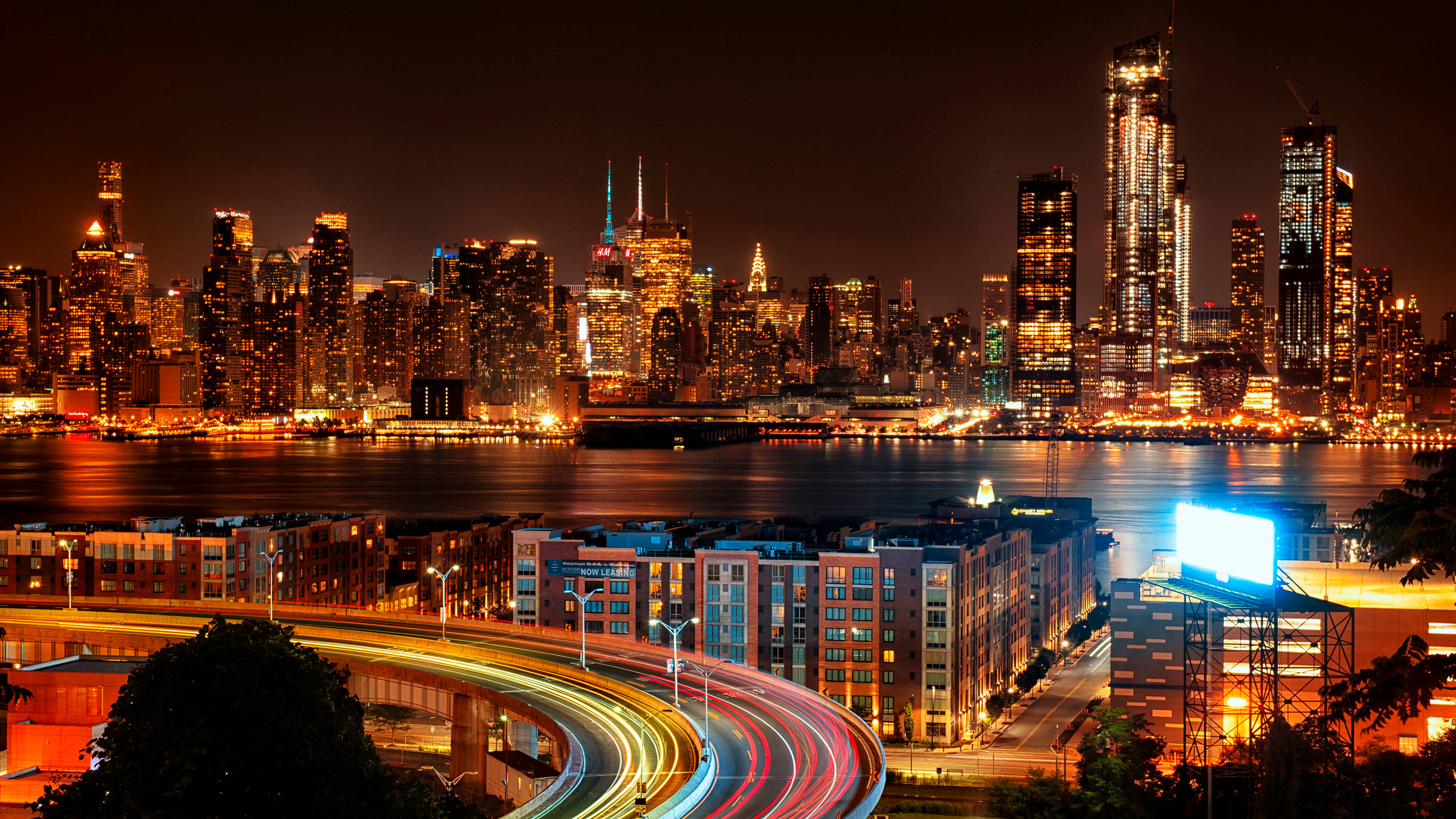 new york 4K 桌布 壁紙 — 紐約令人目眩的夜景，閃爍的摩天大樓和彎曲高速公路上車流形成的鮮豔光軌。