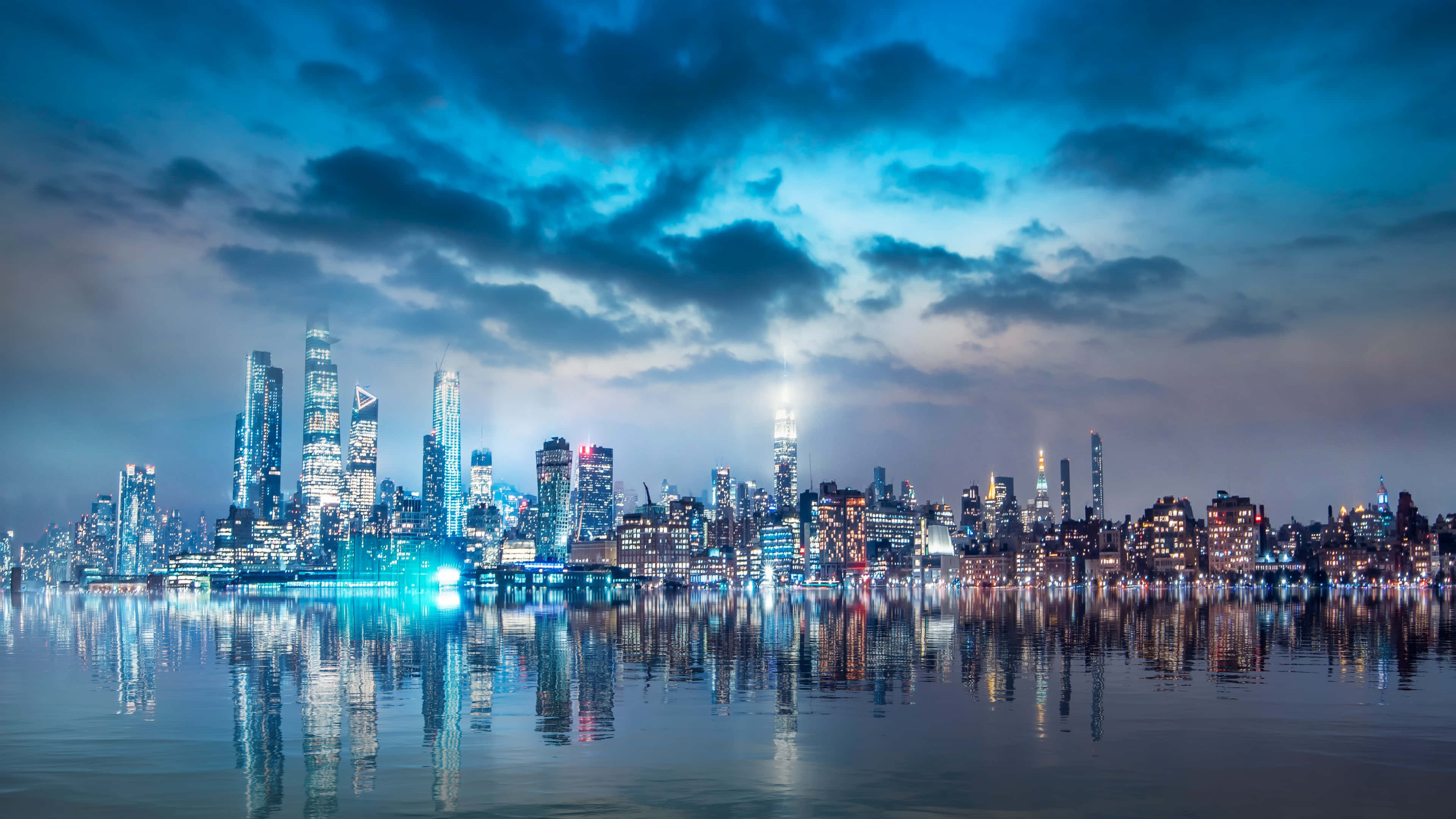 城市景觀 桌布 壁紙 — 紐約令人驚嘆的夜景，摩天大樓在戲劇性的藍天下閃耀，倒映在平靜的水面上。