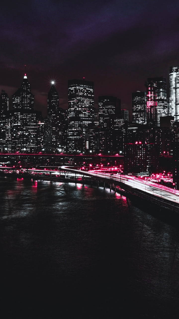 夜間城市天際線，閃爍的建築物和粉紅色燈光的橋樑在水面上。