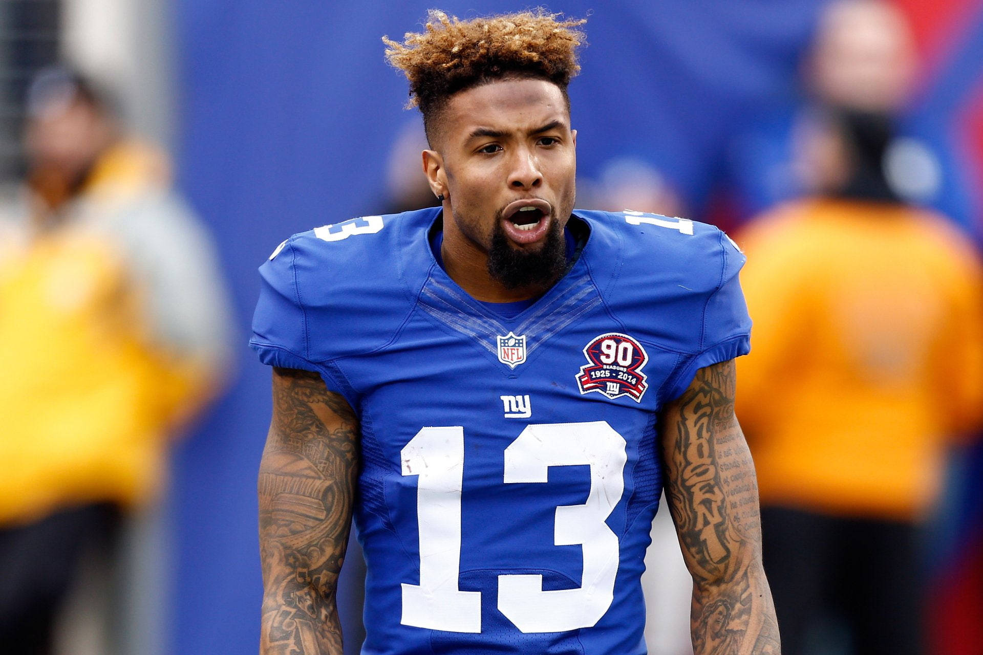 紐約巨人隊的威嚇 壁紙 — 一名專注的NFL球員身穿藍色13號球衣，在模糊的體育場背景下比賽時大喊。