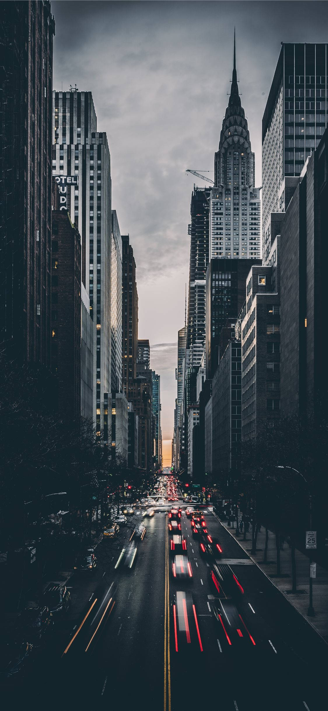 紐約市 iPhone X 行車繁忙街景 壁紙 — 一張長時間曝光照片，拍攝黃昏時分繁忙的城市街道，車燈形成光軌，高聳的摩天大樓在陰雲密佈的天空下。