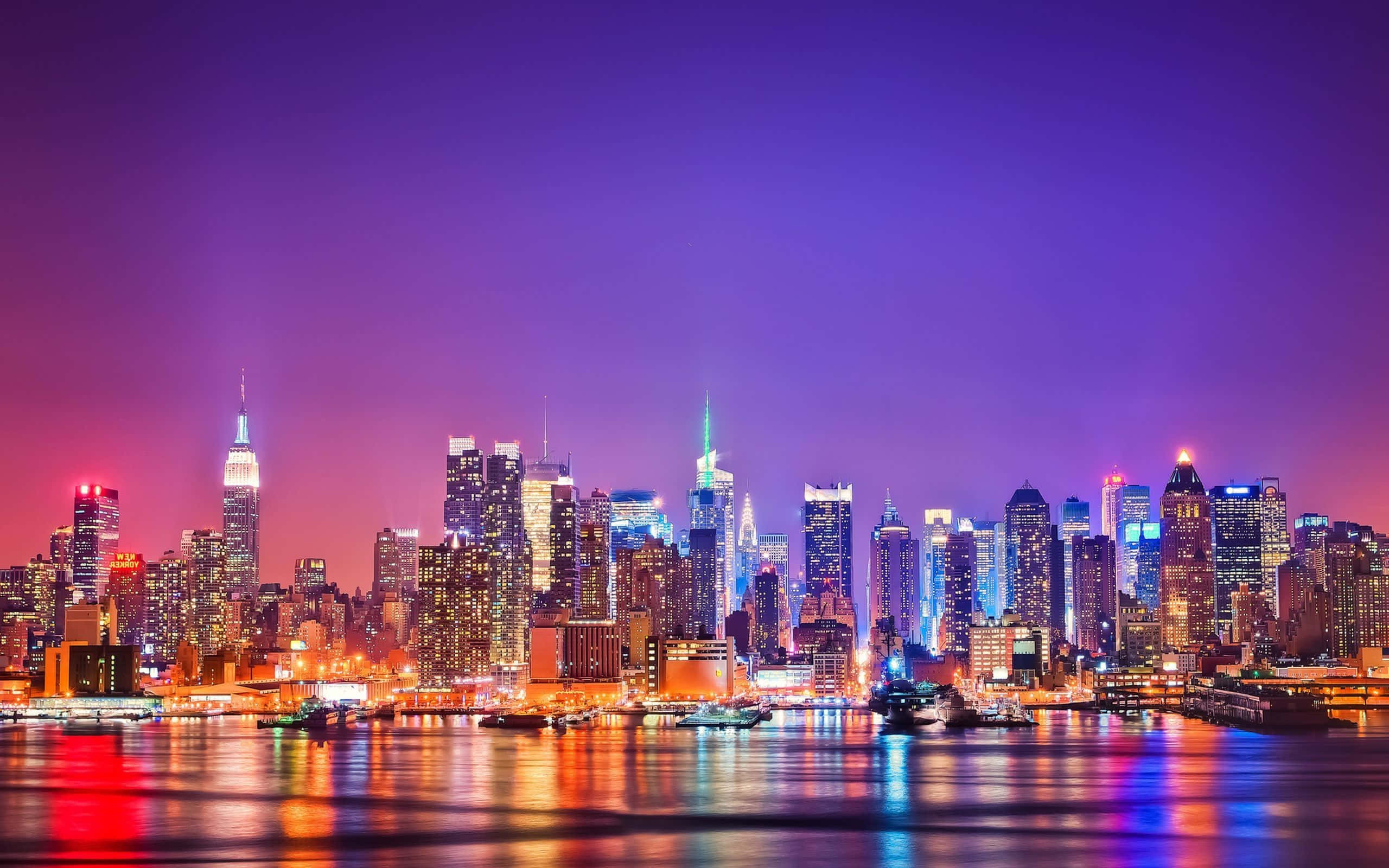 City Lights 桌布 壁紙 — 紐約市充滿活力的夜景，紫色天空下，發光的摩天大樓倒映在水中。