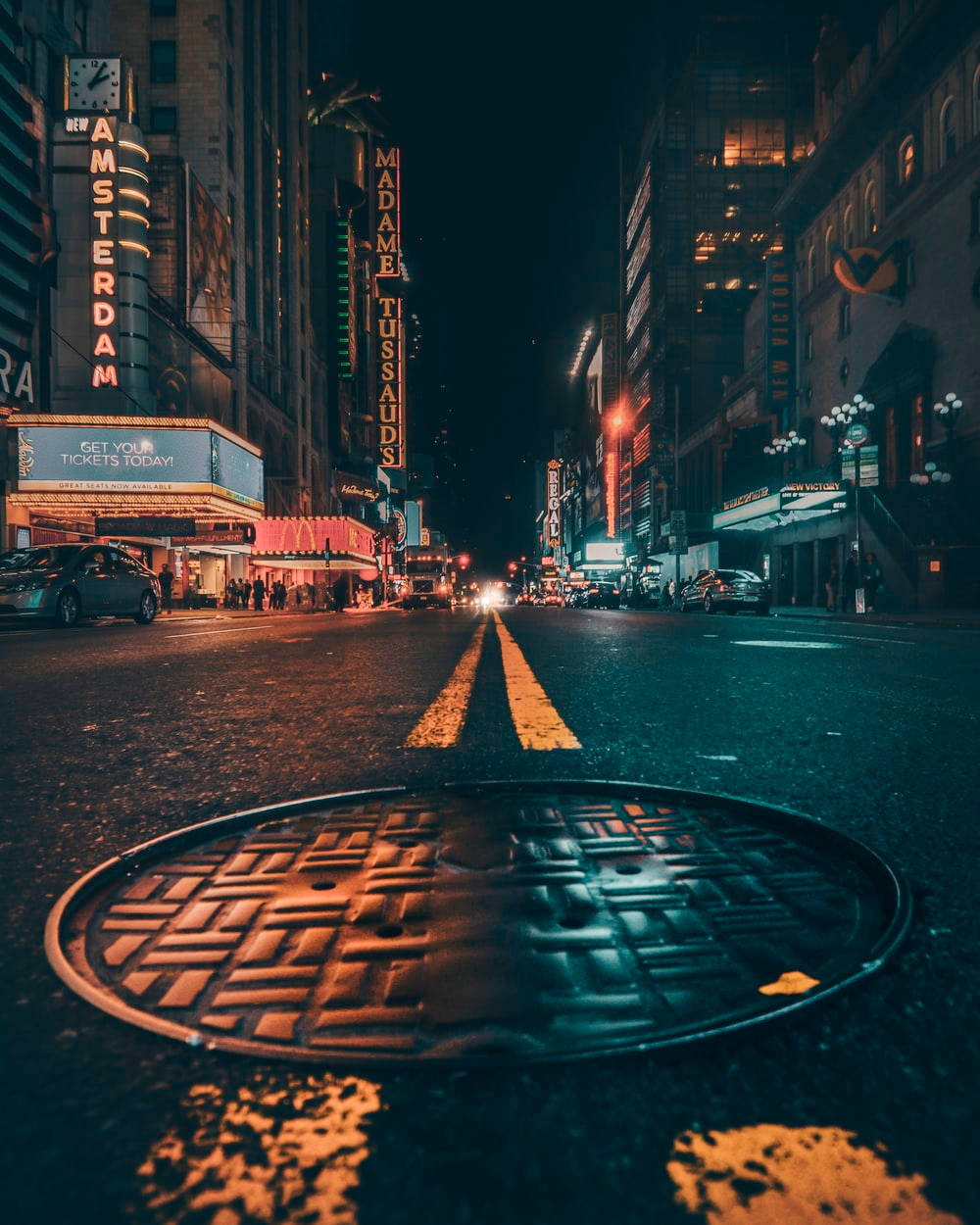 紐約市夜間人行道 壁紙 — 一張低角度夜拍，濕漉漉的城市街道，前景是沙井蓋，還有來自杜莎夫人蠟像館和阿姆斯特丹劇院的霓虹燈招牌。