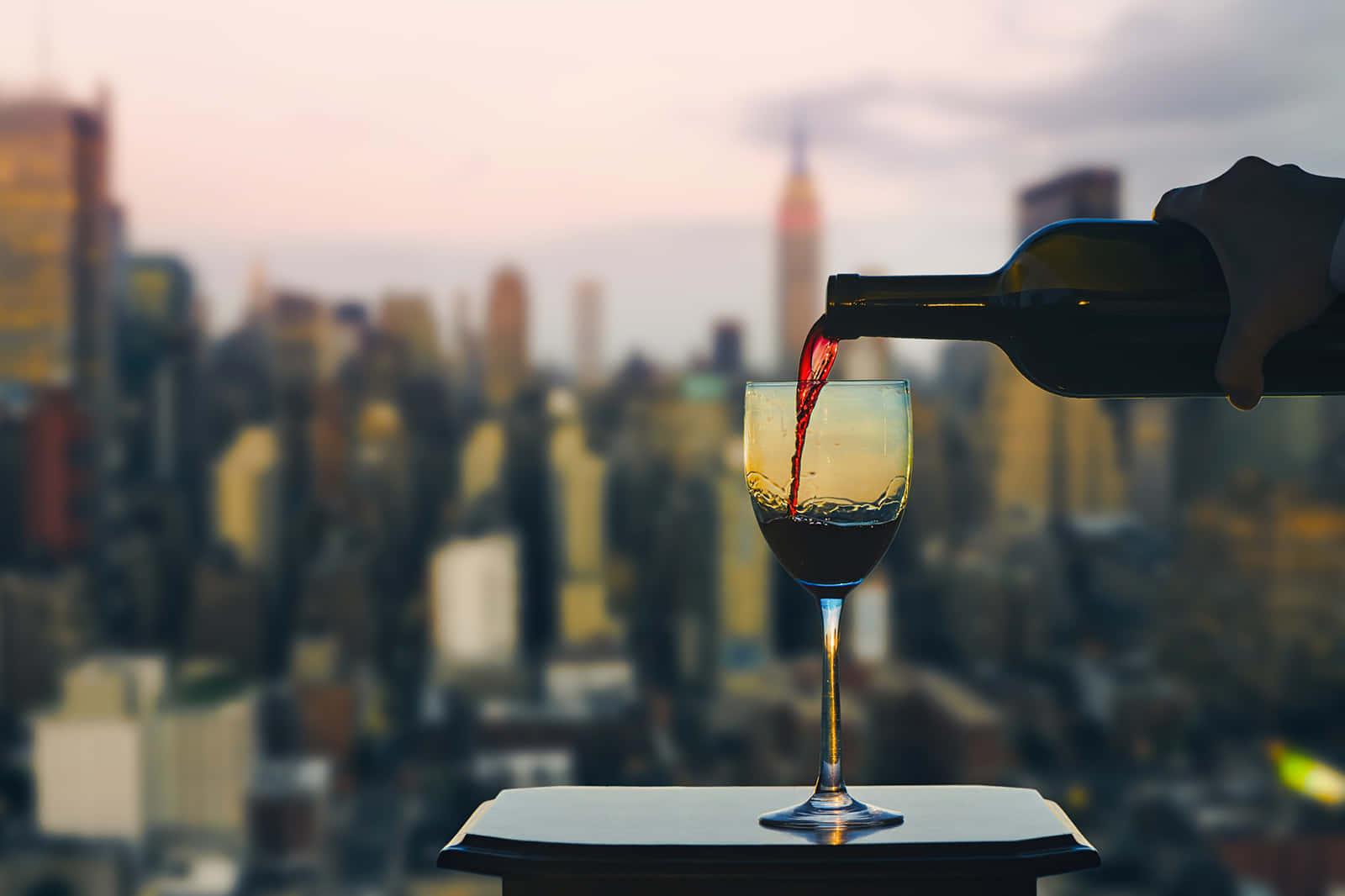 紐約市天際線 壁紙 — 一隻手在黃昏時分將紅酒倒入玻璃杯，背景是模糊的城市天際線。