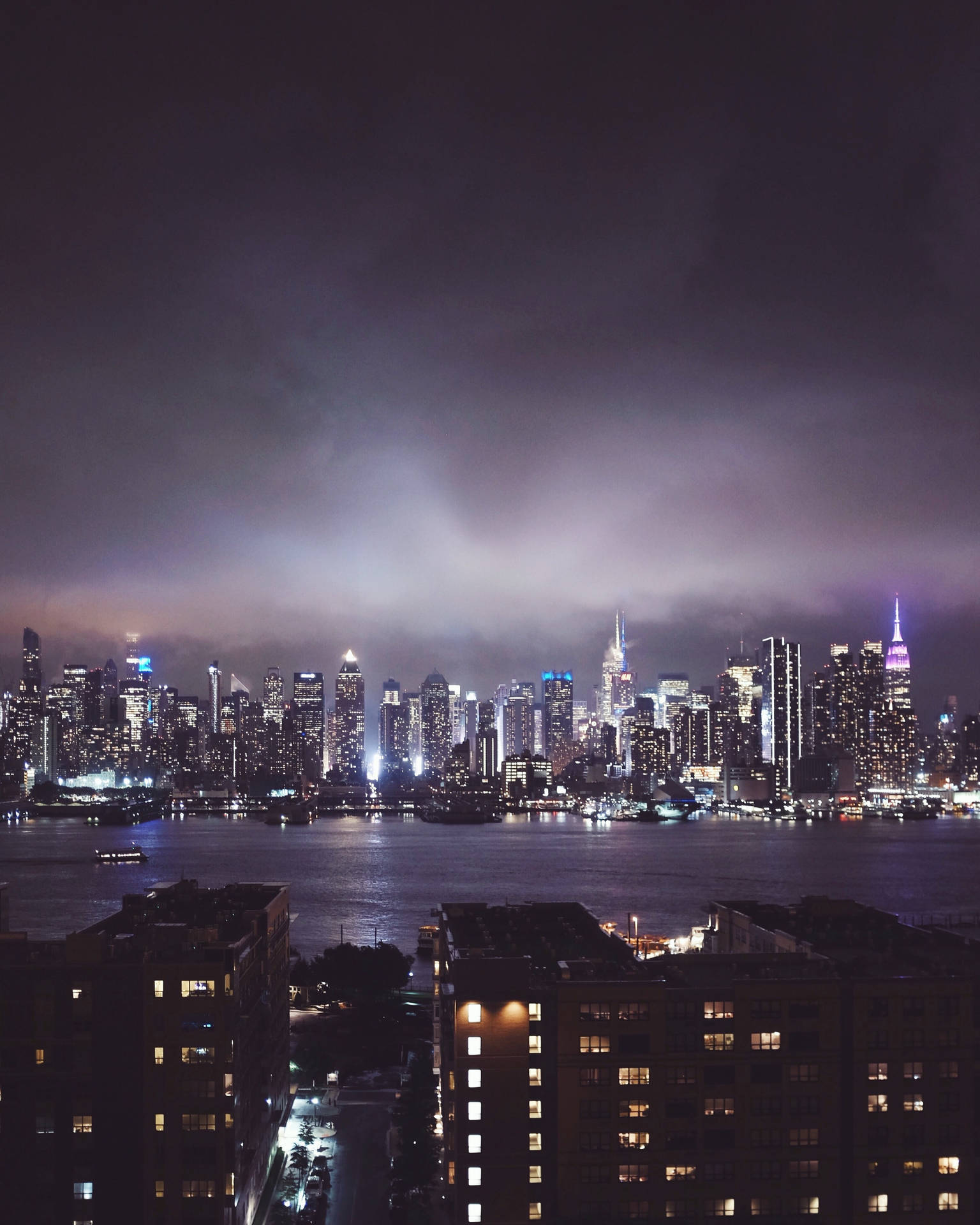 陰天之下，發光的摩天大樓與水面倒影的夜間城市景觀。