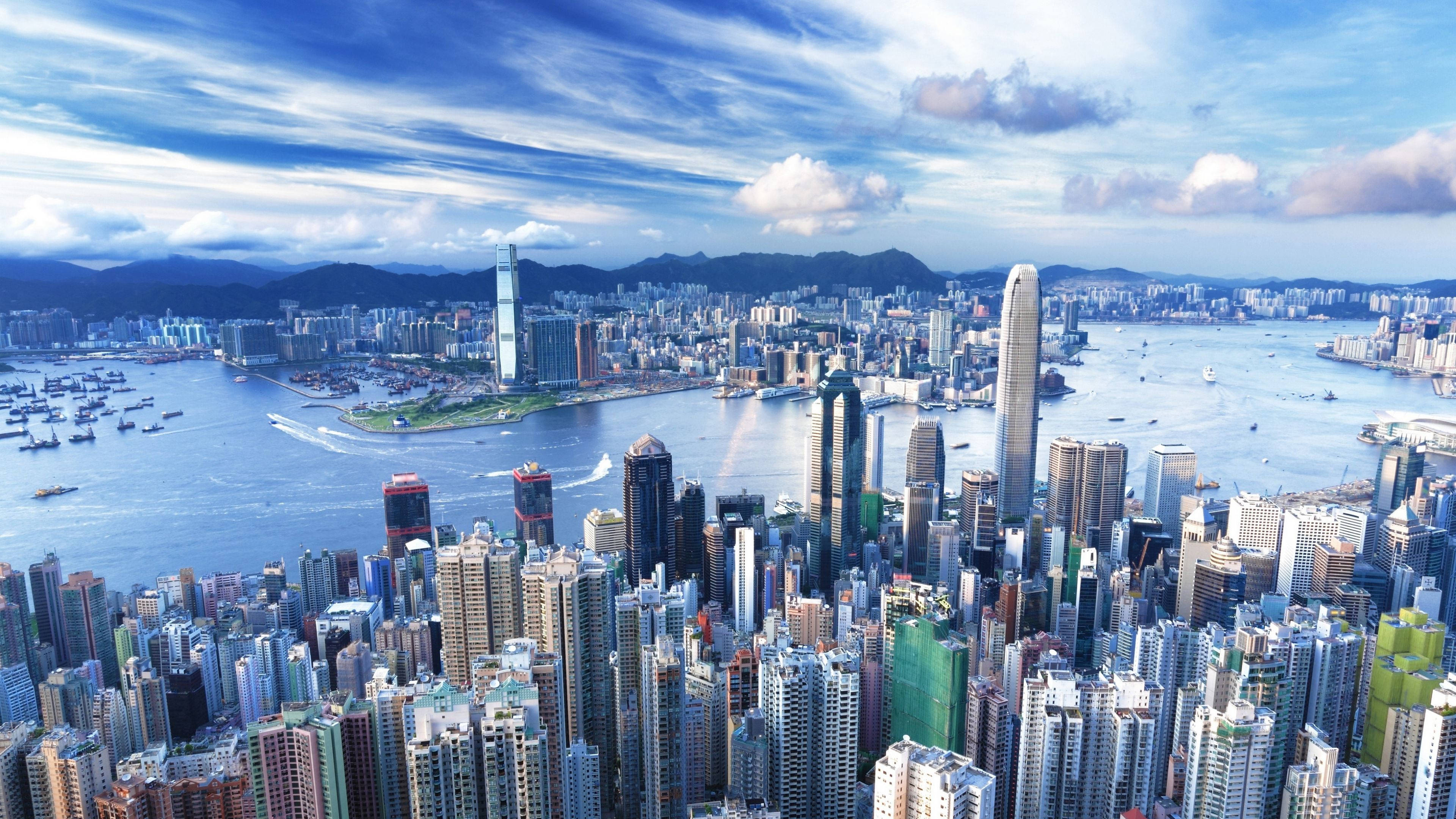 紐約市的壯麗城市景觀 壁紙 — 香港密集天際線的鳥瞰圖，高聳的摩天大樓、維多利亞港和遠處的山脈，在明亮的藍天白雲下。