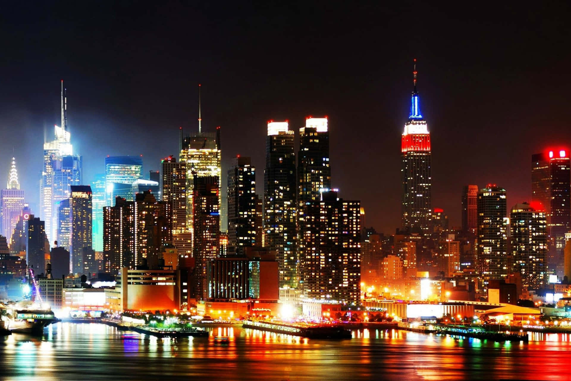 紐約市的心臟 壁紙 — 紐約令人驚嘆的夜景，摩天大樓在黑暗的天空中閃耀， vibrant 燈光在水面上反射。