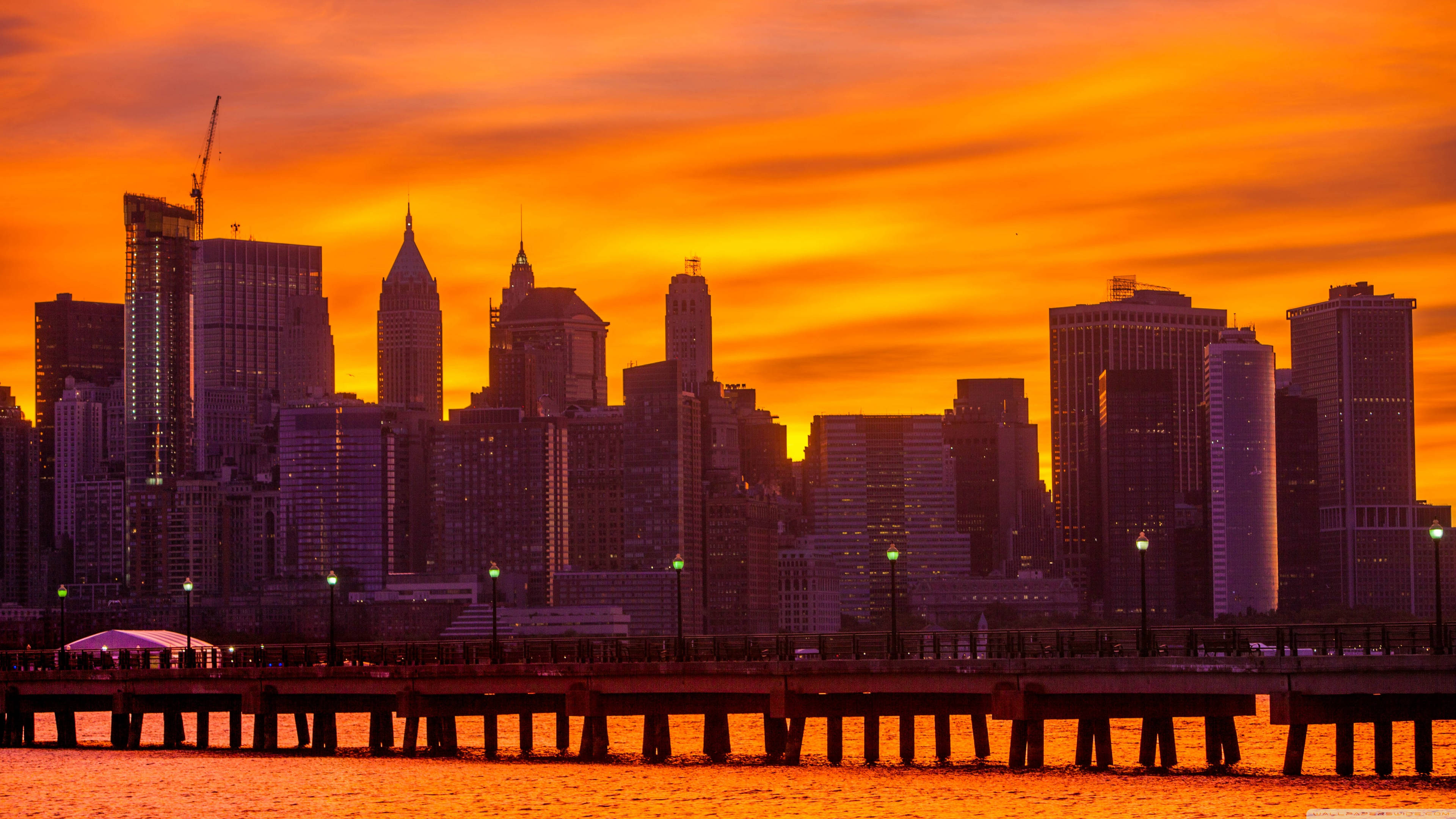 紐約市的絕美風景 壁紙 — 鮮豔的橙色夕陽將紐約市天際線沐浴在溫暖的光芒中，剪影般的摩天大樓矗立在海濱碼頭之上。