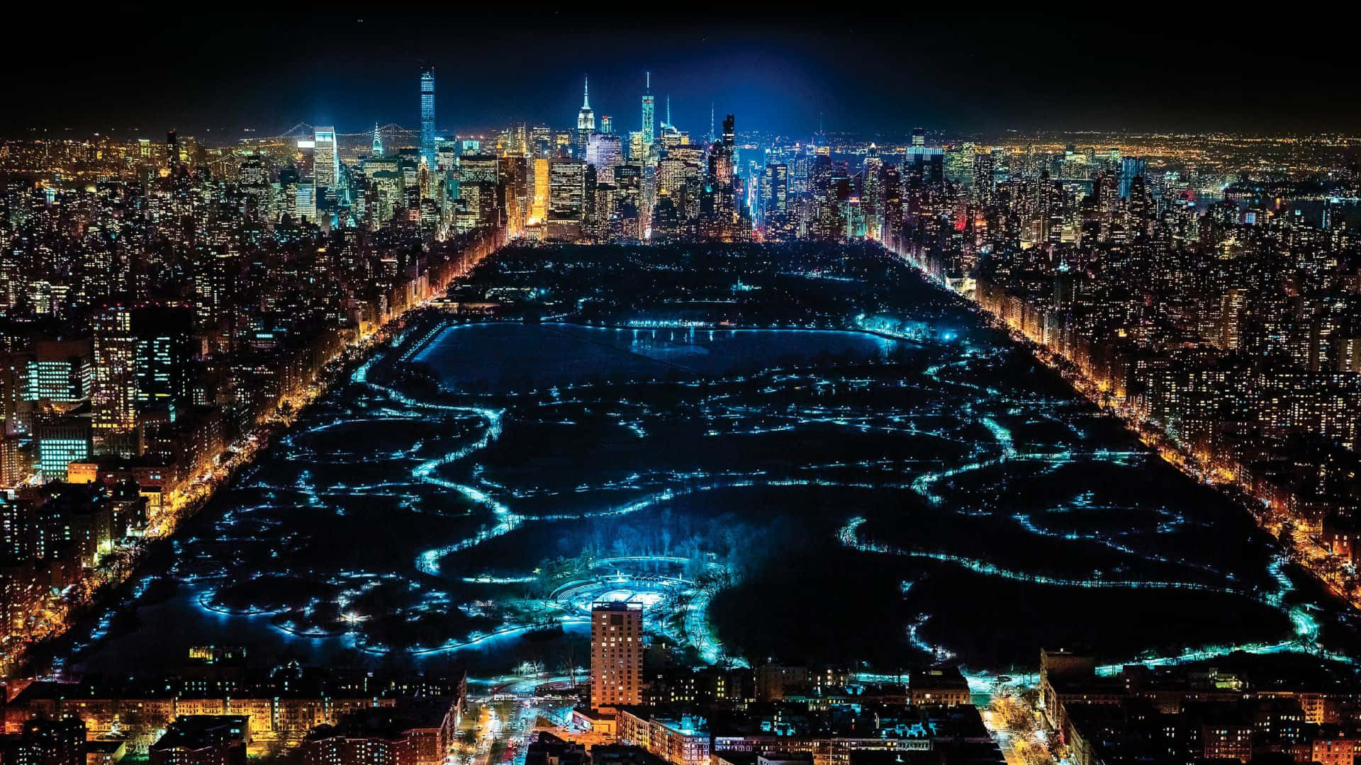 紐約市的美麗 壁紙 — 紐約市中央公園的夜間空中景觀，藍色燈光蜿蜒穿過其小徑和湖泊，周圍環繞著燈火通明的摩天大樓。