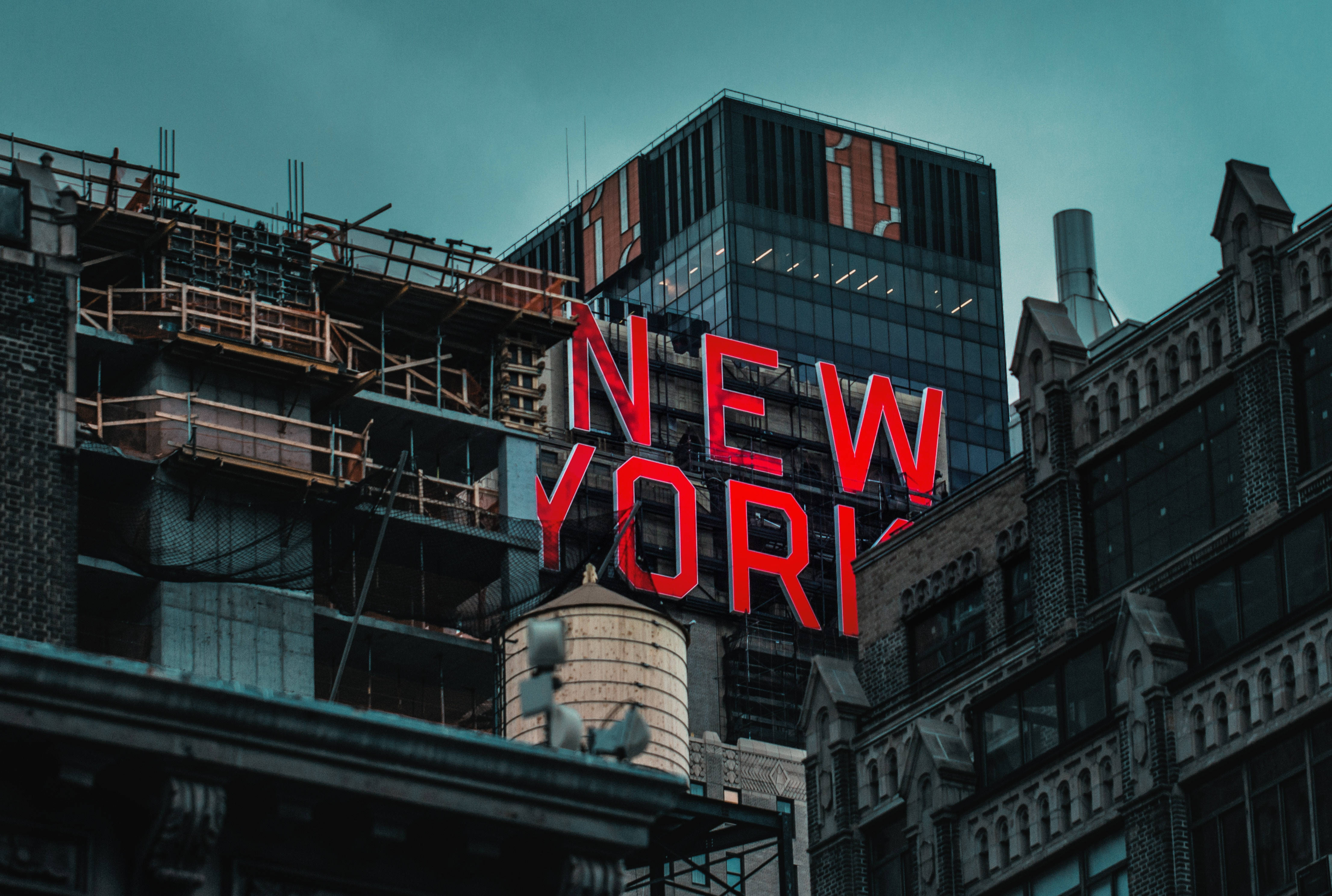 紐約市紅色霓虹招牌建築 壁紙 — 一個大膽的紅色「NEW YORK」標誌在憂鬱的藍綠色天空下發光，下方是歷史磚砌建築和現代玻璃塔樓的混合體，正在建設中。