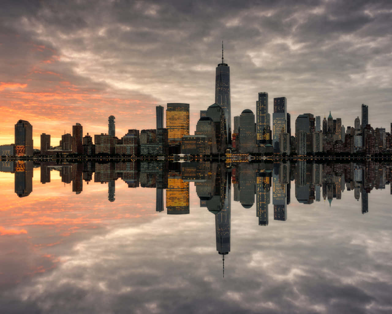 紐約市絢麗的夜景 壁紙 — 紐約市天際線的壯麗日落，摩天大樓在平靜的水面上完美倒映，天空佈滿戲劇性的橙色和灰色雲彩。