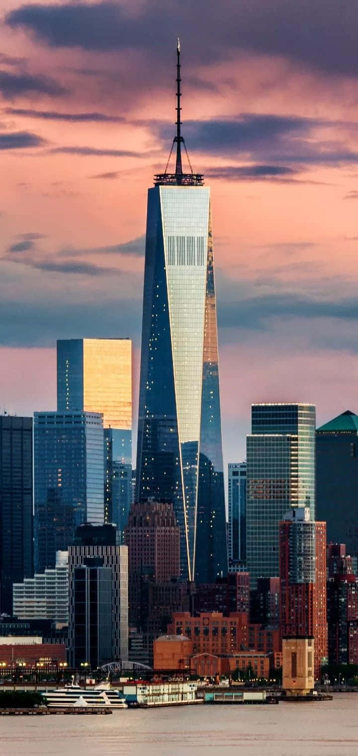 日落時分的世界貿易中心一號大樓，粉紅色的雲彩和城市燈光倒映在水面上。