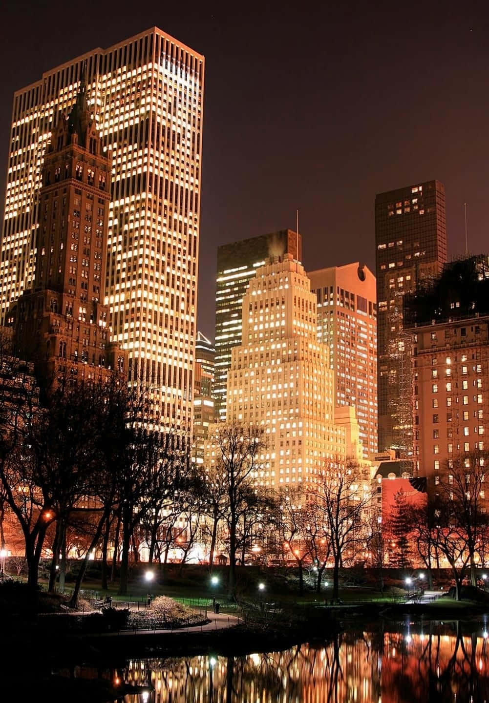 紐約時尚風景概覽 壁紙 — 高聳的城市摩天大樓在夜晚溫暖地發光，它們的燈光倒映在一個被光禿樹木環繞的寧靜池塘中。