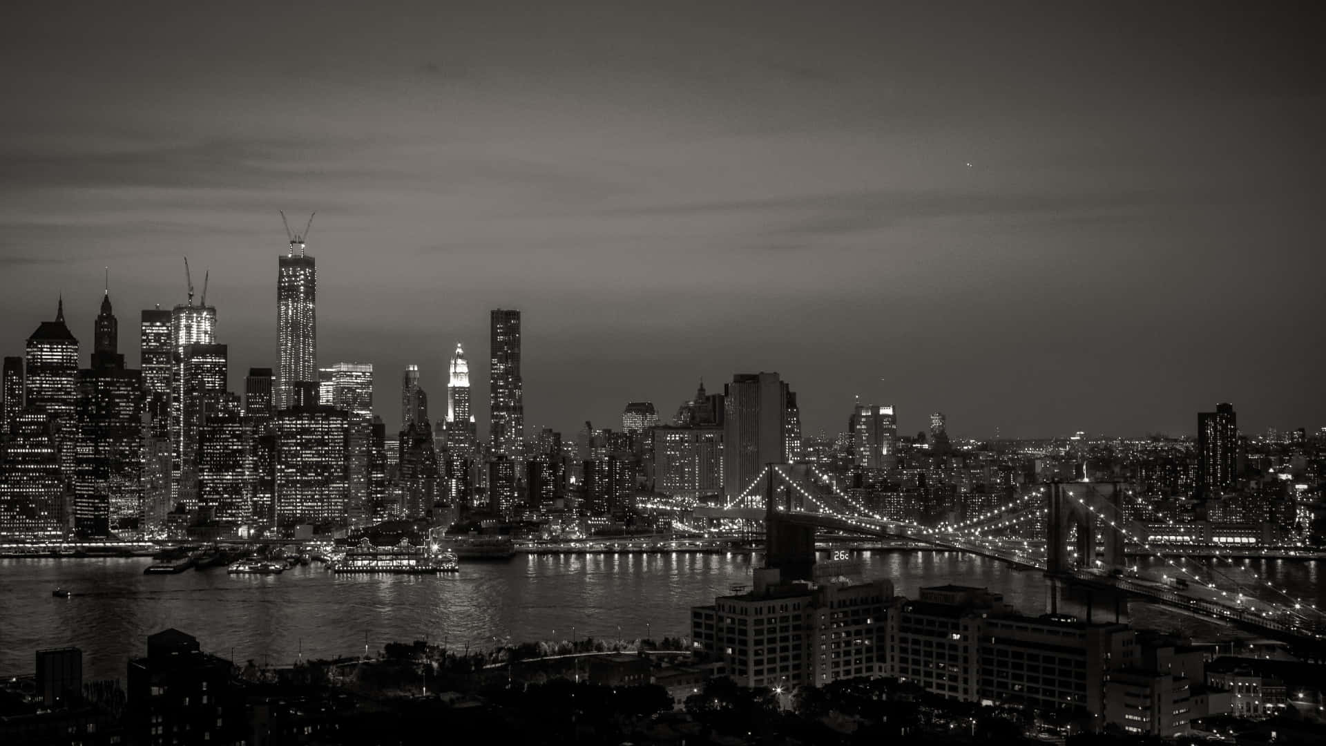 Black And White Cityscape 桌布 壁紙 — 紐約的單色夜景，有燈火通明的摩天大樓和一座在水面上亮燈的橋樑。