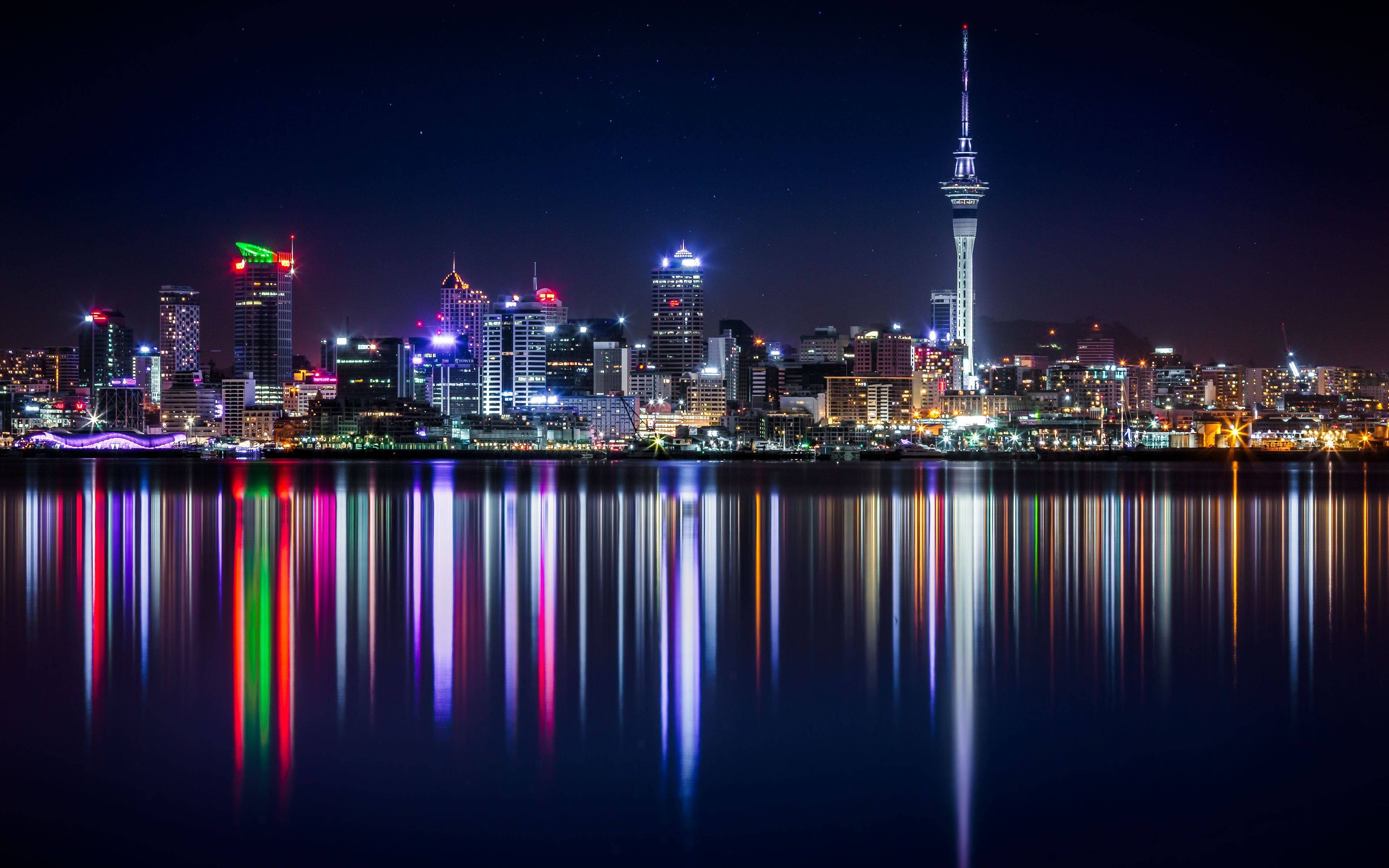 紐西蘭奧克蘭夜景燈光 壁紙 — 充滿活力的城市天際線夜景，色彩繽紛的燈光在星空下平靜的水面上完美倒映。