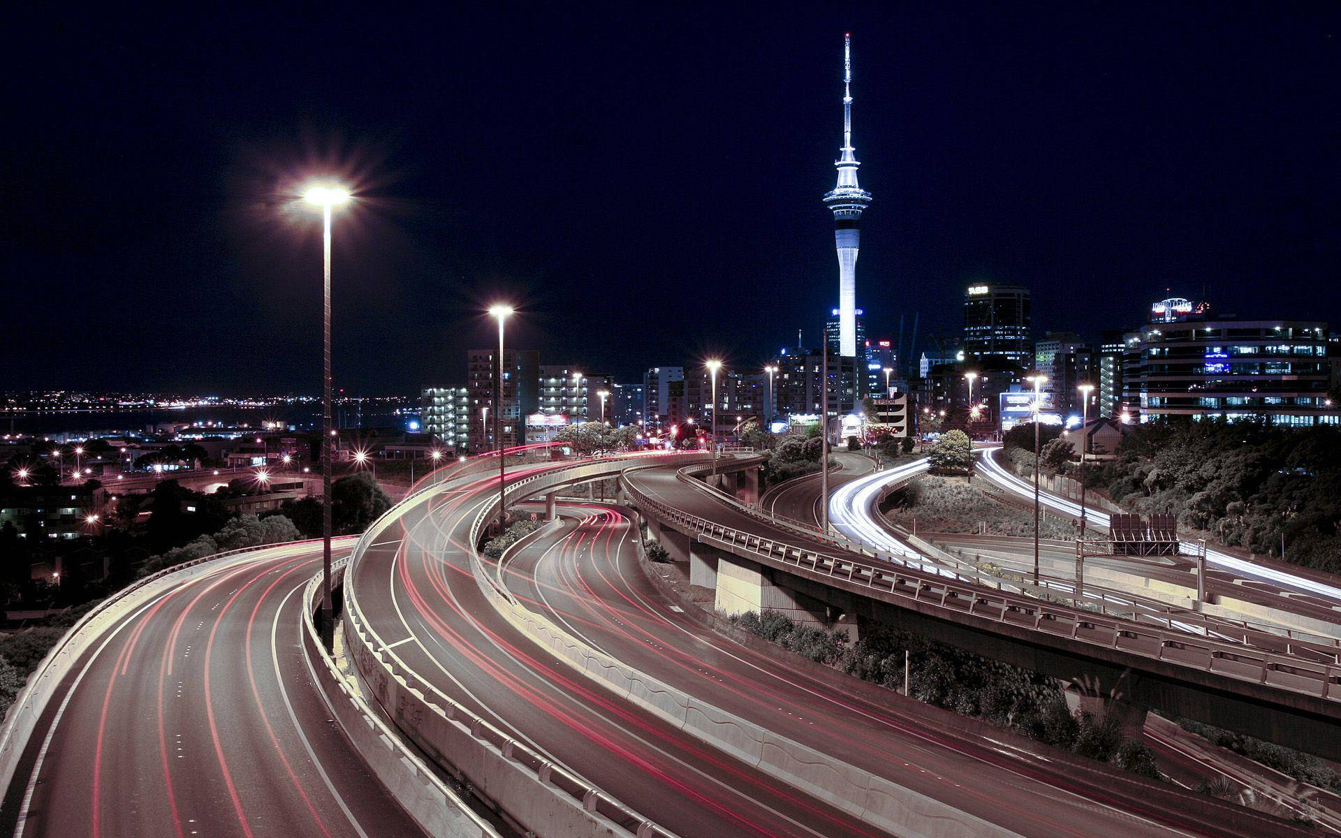 紐西蘭奧克蘭市壯麗景色 壁紙 — 充滿活力的夜景城市景觀，特色是發光的高速公路、流動的車燈，以及一座高聳的照明摩天大樓，映襯著黑暗的天空。