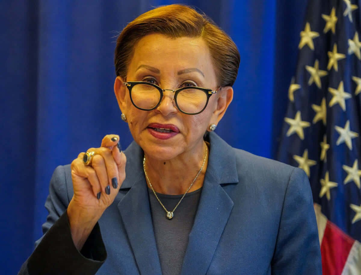 紐迪亞·維拉茲克斯穿藍色服裝與藍色窗簾 壁紙 — 一名身穿藍色西裝、戴眼鏡的女性在藍色窗簾和美國國旗前說話時做手勢。