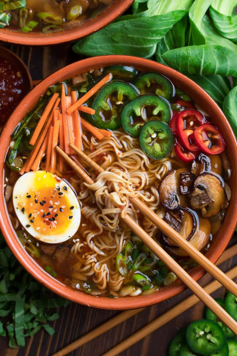 素食拉麵 壁紙 — 一碗熱騰騰的拉麵，上面放著半熟蛋、切片墨西哥辣椒、蘑菇和紅蘿蔔，周圍環繞著新鮮蔬菜。