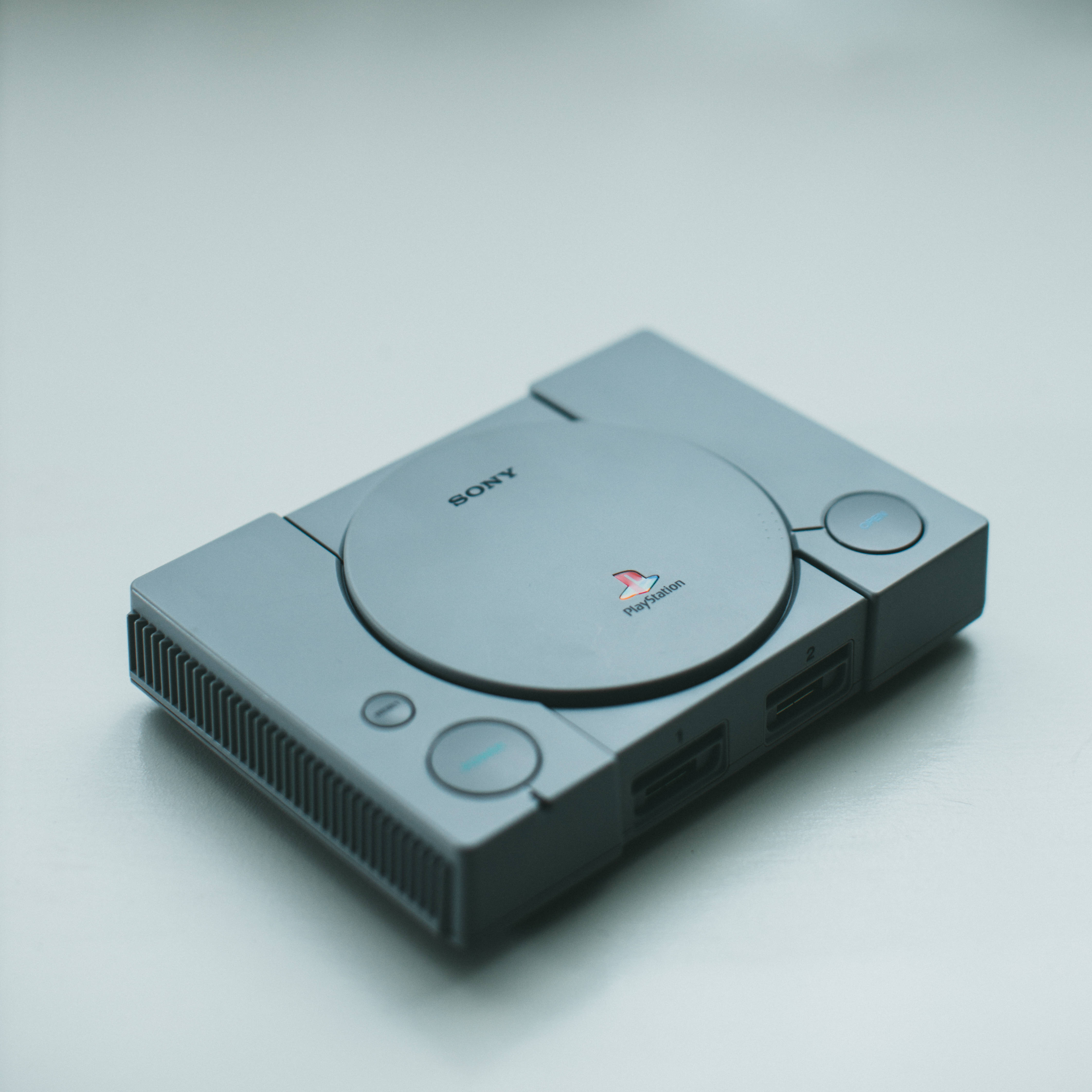 索尼 PlayStation Classic 壁紙 — 一部時尚的銀色 PlayStation 1 主機放置在簡約的白色表面上，柔和的光線突顯其標誌性的圓形設計。