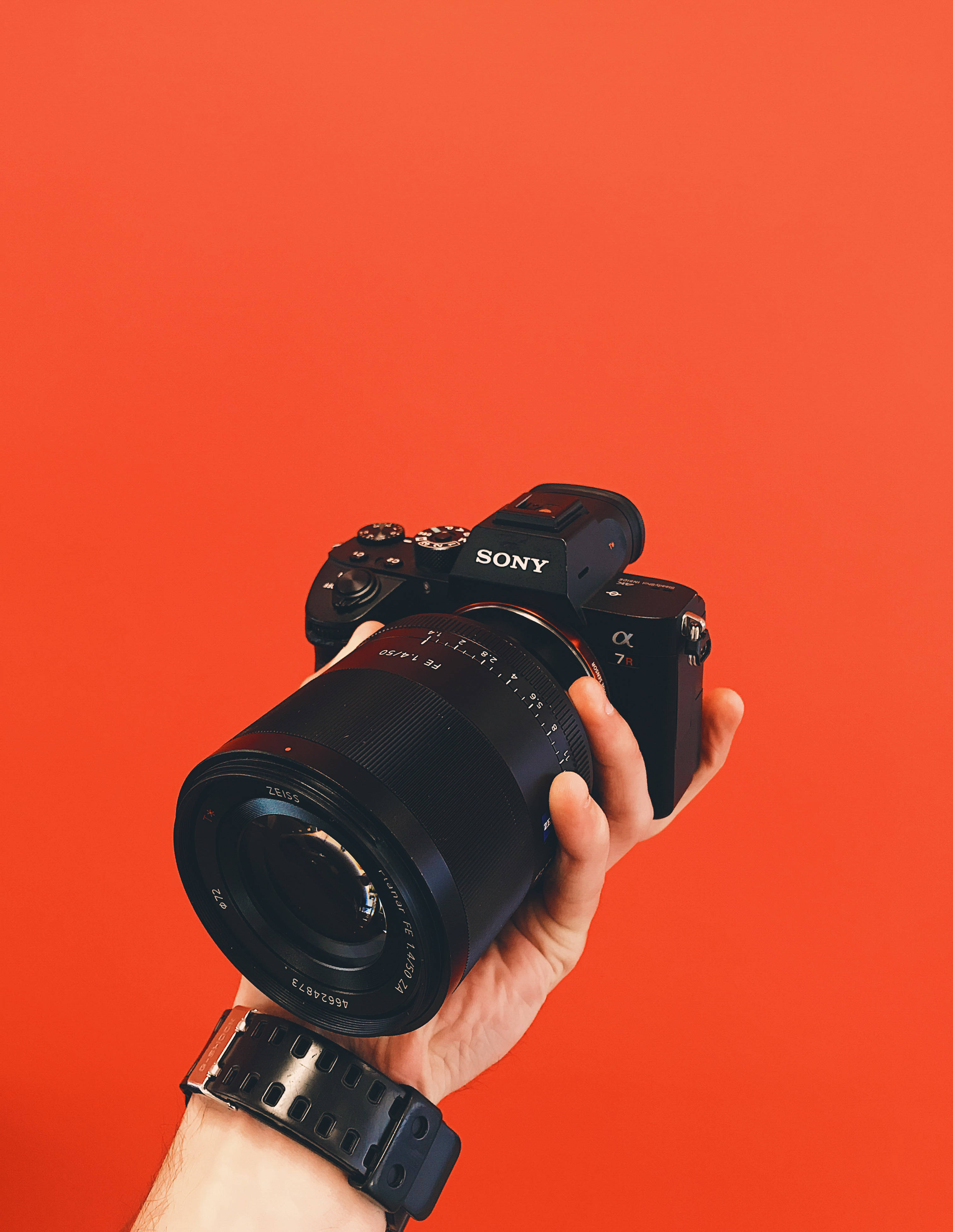索尼Alpha單眼相機 壁紙 — 一隻手拿著黑色Sony Alpha 7R相機，背景為鮮豔的橙色。