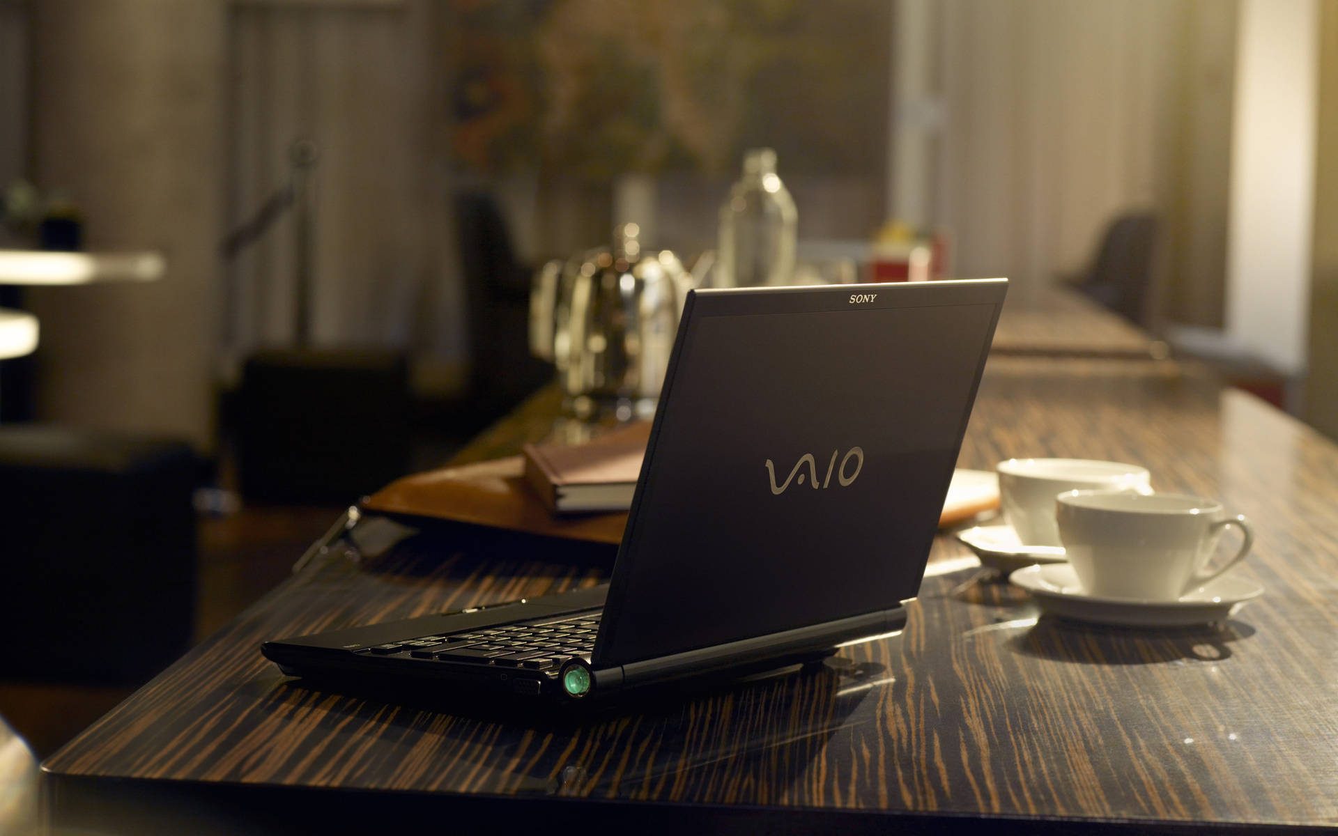 索尼Vaio筆記本電腦 壁紙 — 一部黑色Sony VAIO手提電腦打開放在深色木桌上，旁邊有兩個白色咖啡杯，在一個燈光溫暖的咖啡館裏。