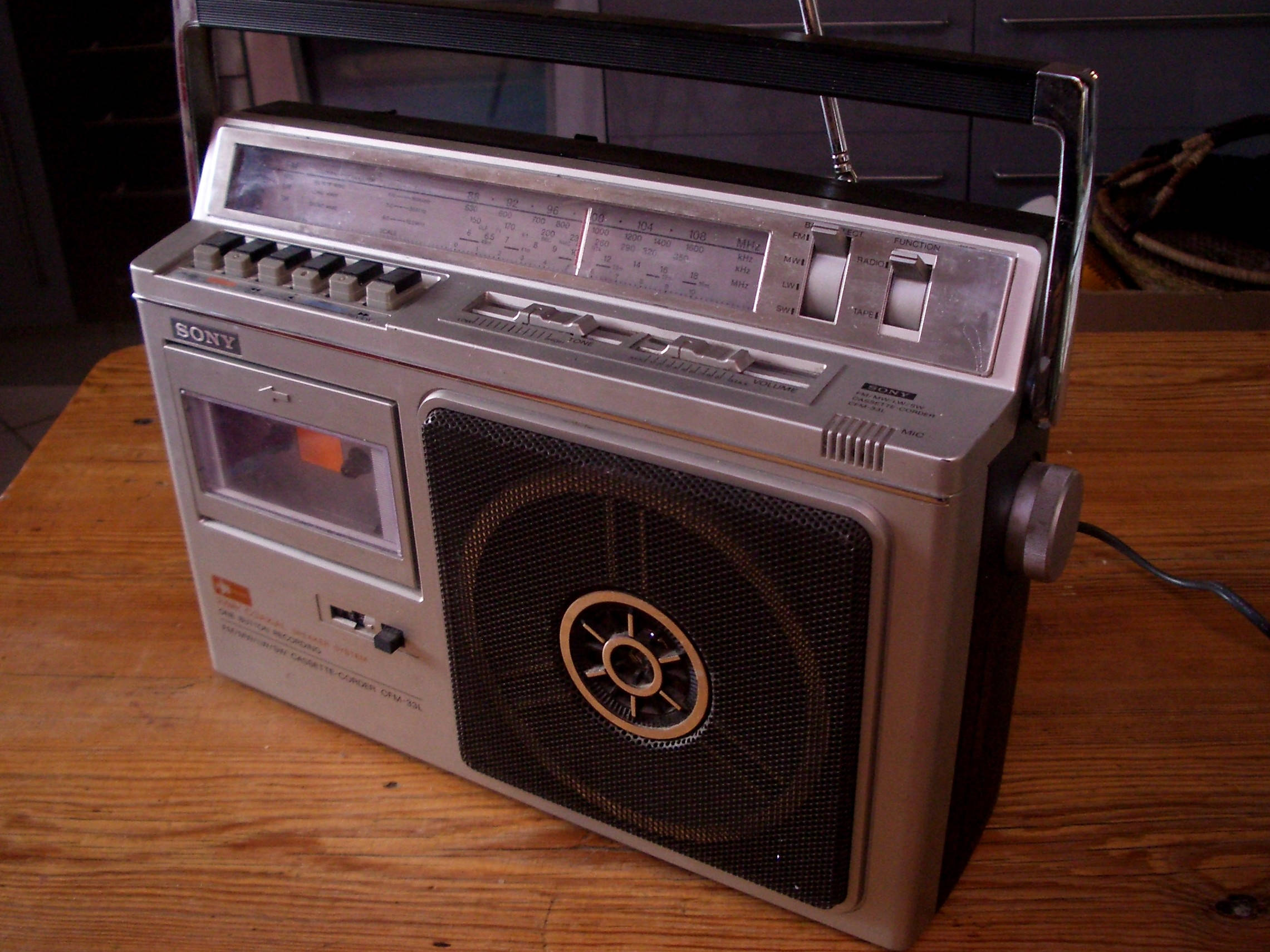 索尼老式錄音帶機 壁紙 — 復古銀色Sony手提音響，帶卡式錄音帶座和大喇叭，放在木桌上。