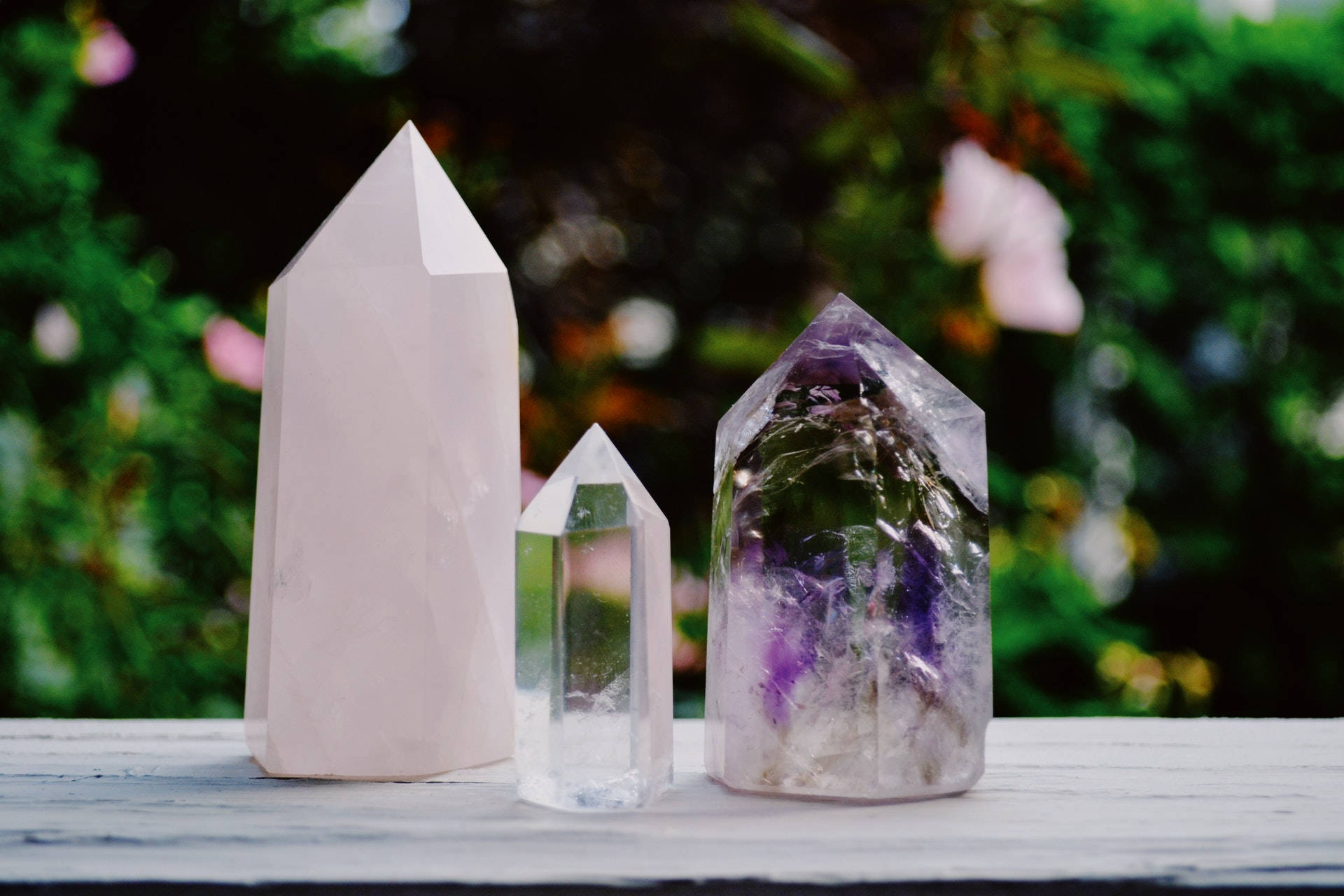 紫水晶單尖塔 壁紙 — 三顆拋光的水晶尖 玫瑰石英 透明石英 紫水晶 放置在風化的木質表面上，背景是柔和模糊的花園。