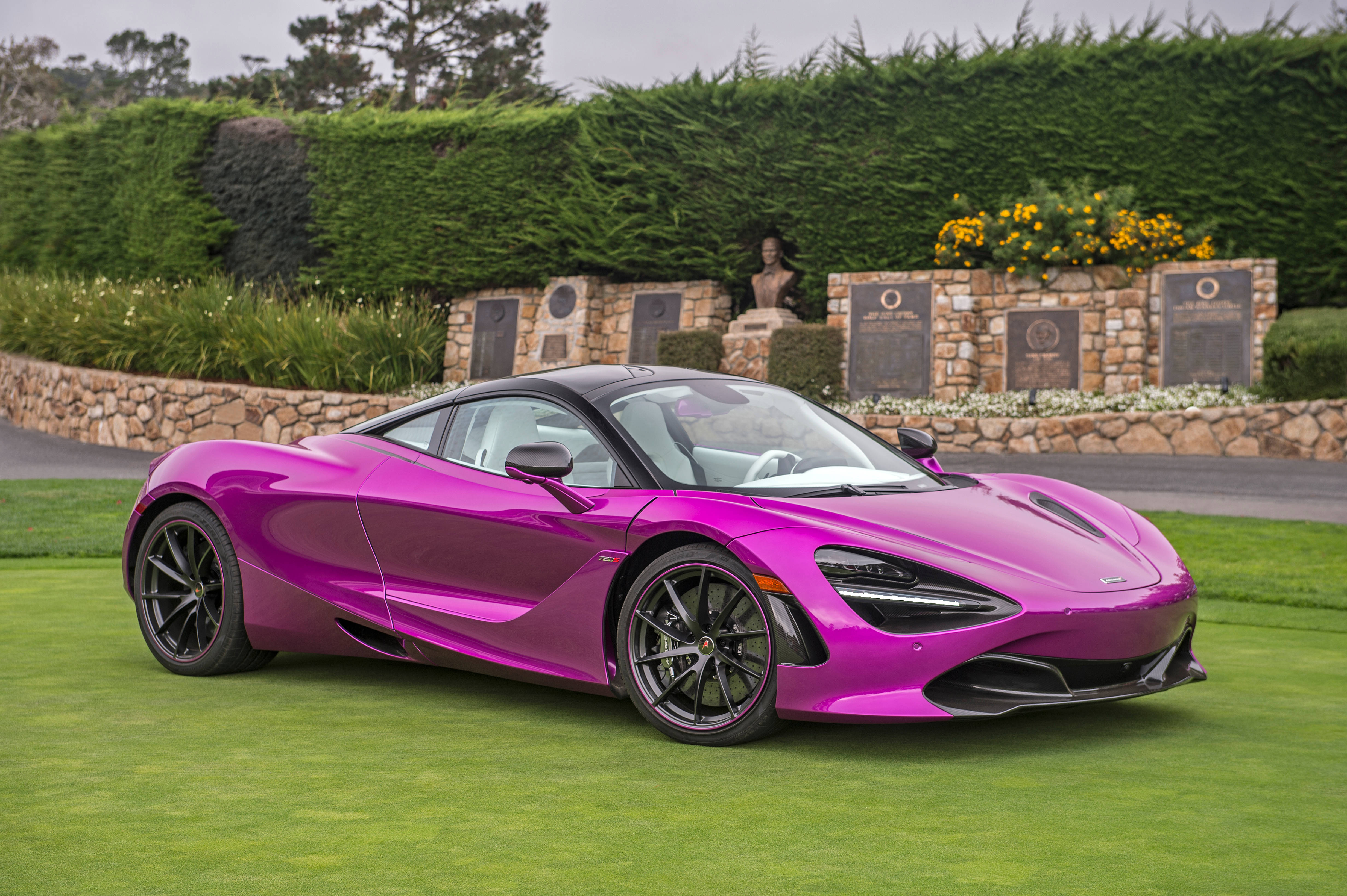 紫紅色麥拉倫720S 壁紙 — 一輛充滿活力的洋紅色McLaren 720S跑車在修剪整齊的綠色草坪上閃閃發光，旁邊是帶有牌匾和黃色花朵的石牆。