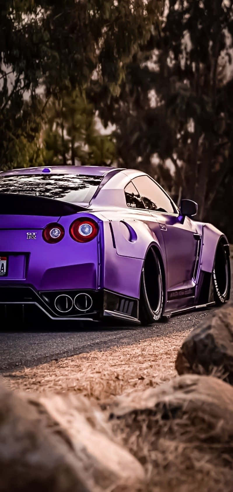紫色 Nissan R35 GT-R 車圖 壁紙 — 一輛充滿活力的紫色日產GT-R跑車停泊在碎石路上，周圍環繞著樹木和岩石。