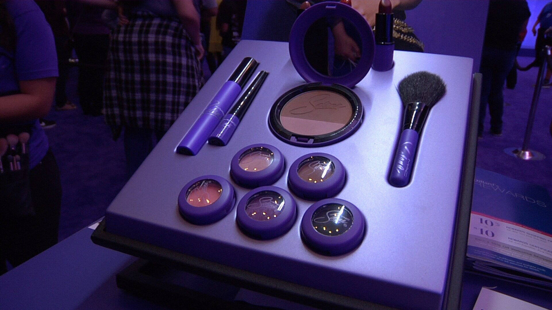 Mac Cosmetics x Selena Quintanilla 壁紙 — 紫色主題化妝品展示，包括粉盒、刷子和鉛筆，排列在會議的亮燈桌上。