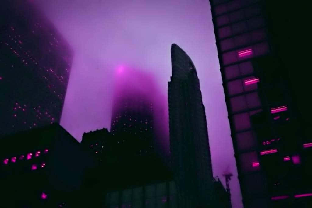 紫色唯美霧氣城市建築畫面 壁紙 — 紫色霓虹燈穿過朦朧的夜空，照亮高聳的摩天大樓，呈現未來主義的城市景觀。