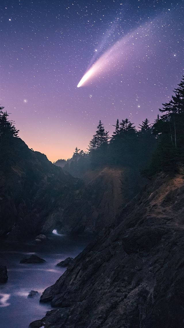 紫色星雲iPhone流星壁紙 壁紙 — 一顆明亮的彗星劃過佈滿星星的黃昏天空，下方是崎嶇的海岸峽谷，有流動的水。